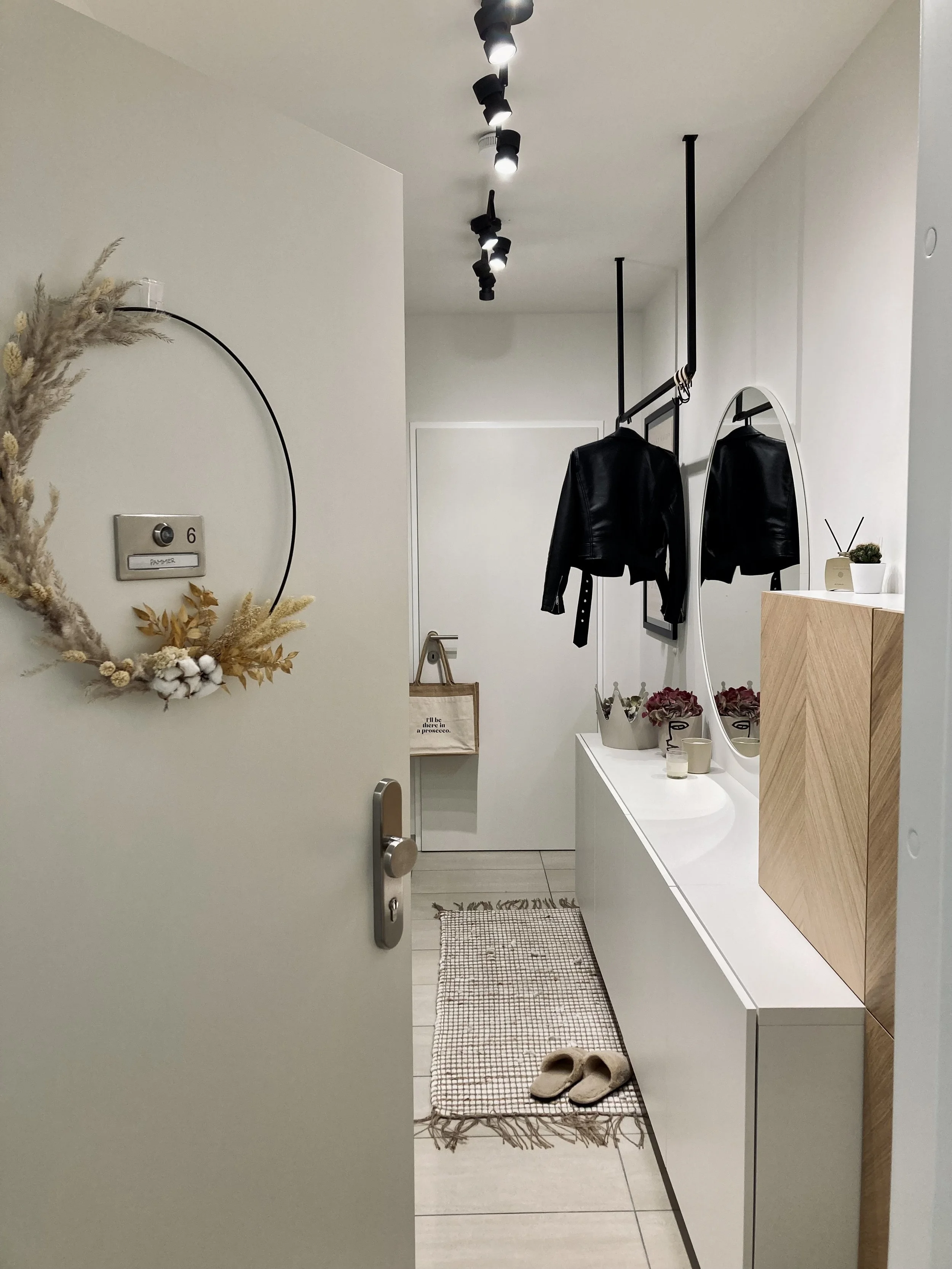 Ein Flur mit weißen Wänden und einem weißen Boden. An der Wand hängt ein runder Dekorkranz mit getrockneten Pflanzen. Auf einem weißen Schrank stehen einige Dekogegenstände, darunter Töpfe mit Blumen, eine kleine Schale und ein Kerzenhalter. Über dem Schrank hängt ein runder Spiegel. An der Wand sind schwarze Kleiderhaken mit einem schwarzen Lederjacke. Auf dem Boden liegt ein kleiner Teppich mit Fransen und bequeme Hausschuhe.