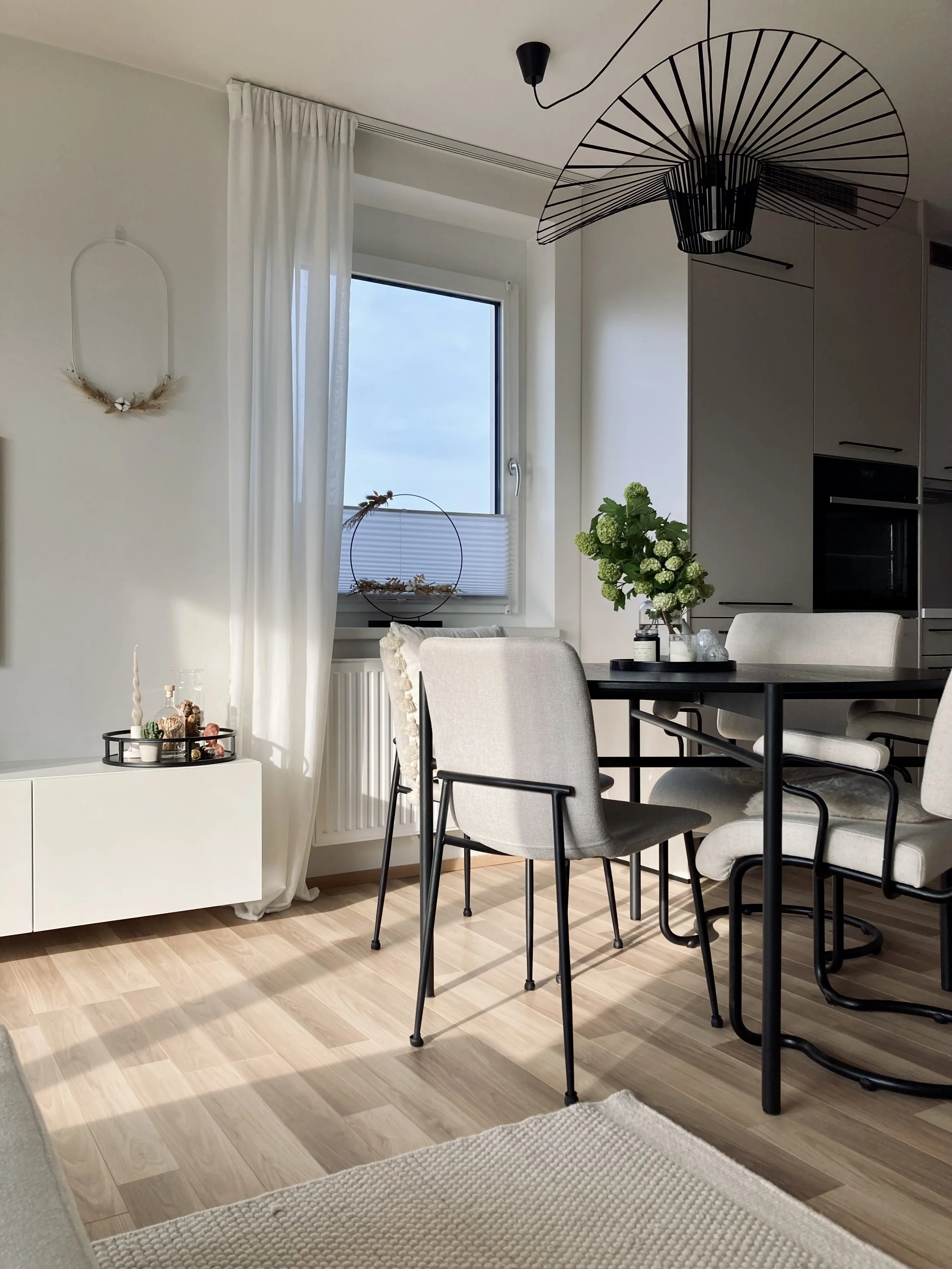 Modernes Esszimmer mit hellen Wänden, einem großen Fenster mit weißen Vorhängen, einem schwarzen Esstisch mit beige Polsterstühlen und einer Vase mit grünen Blumen.