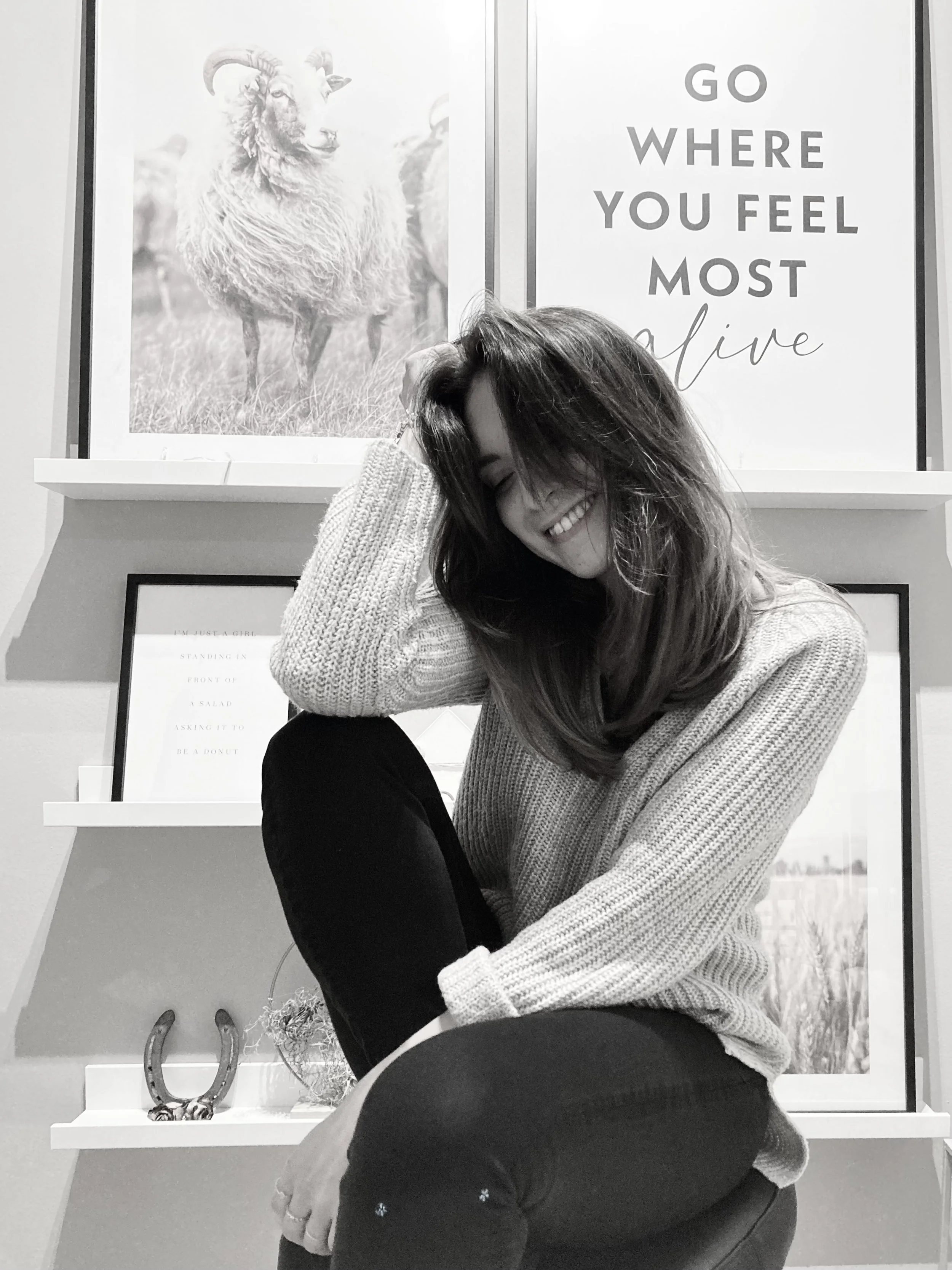 Lächelnde junge Frau in einem Pullover, die auf einem Stuhl sitzt, mit Hintergrundbildern an der Wand, darunter ein Zitat und ein Bild eines Schafes.