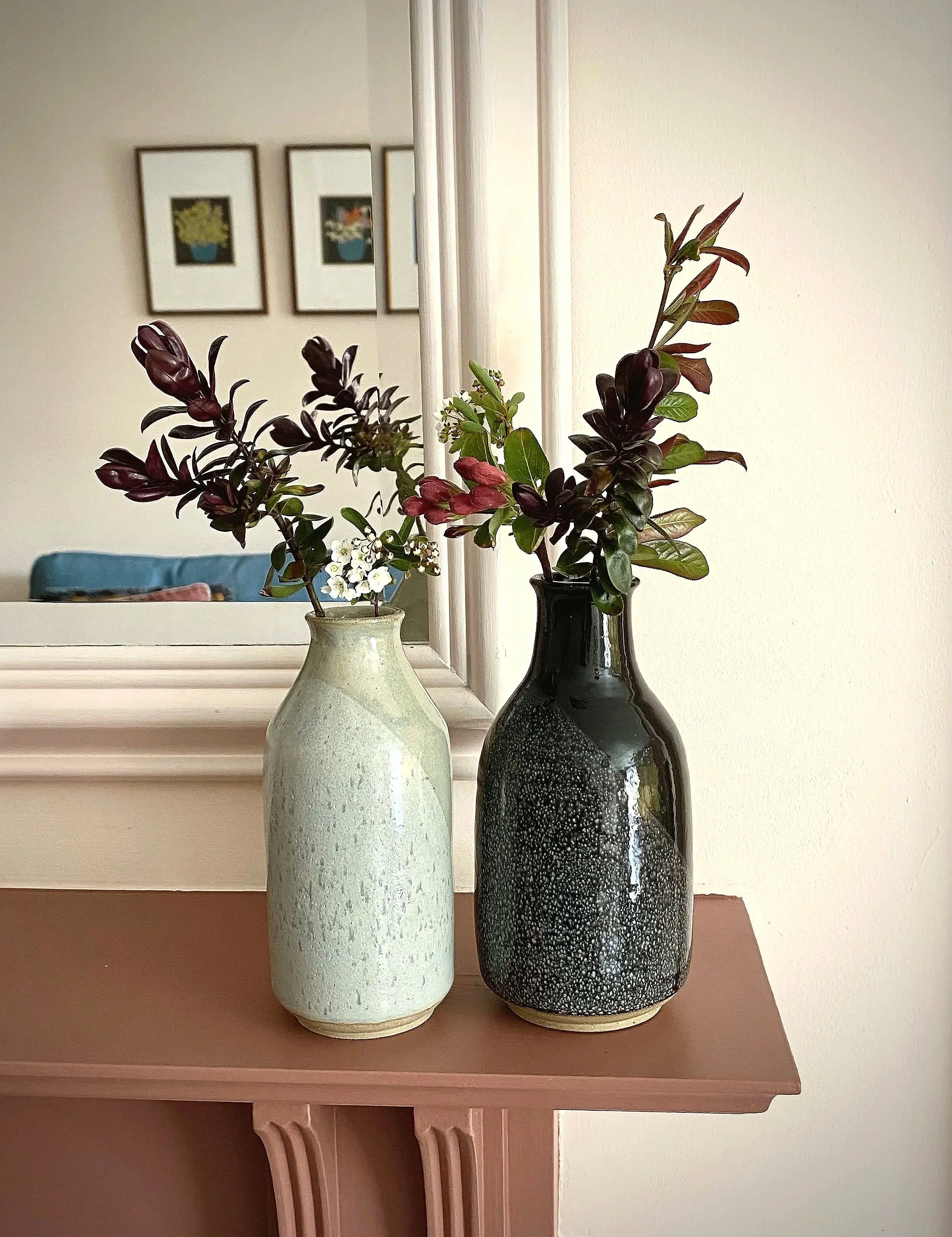 Two vases with leafy branches and small white flowers, placed on a brown wooden mantel in front of a mirror, with framed pictures of plants on a wall reflected in the mirror.