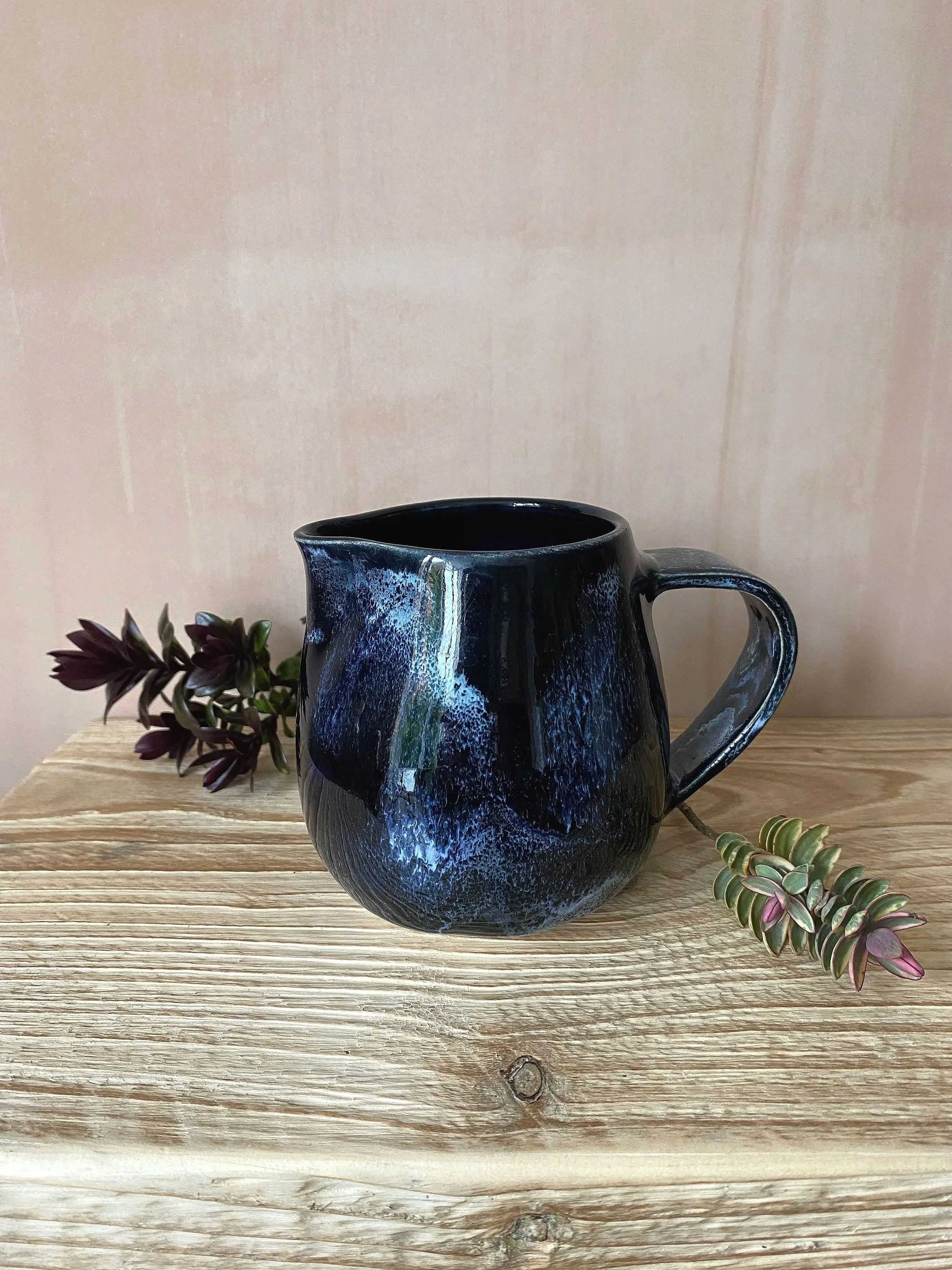 A dark blue ceramic pitcher with a textured surface, placed on a wooden table, with some succulents.
