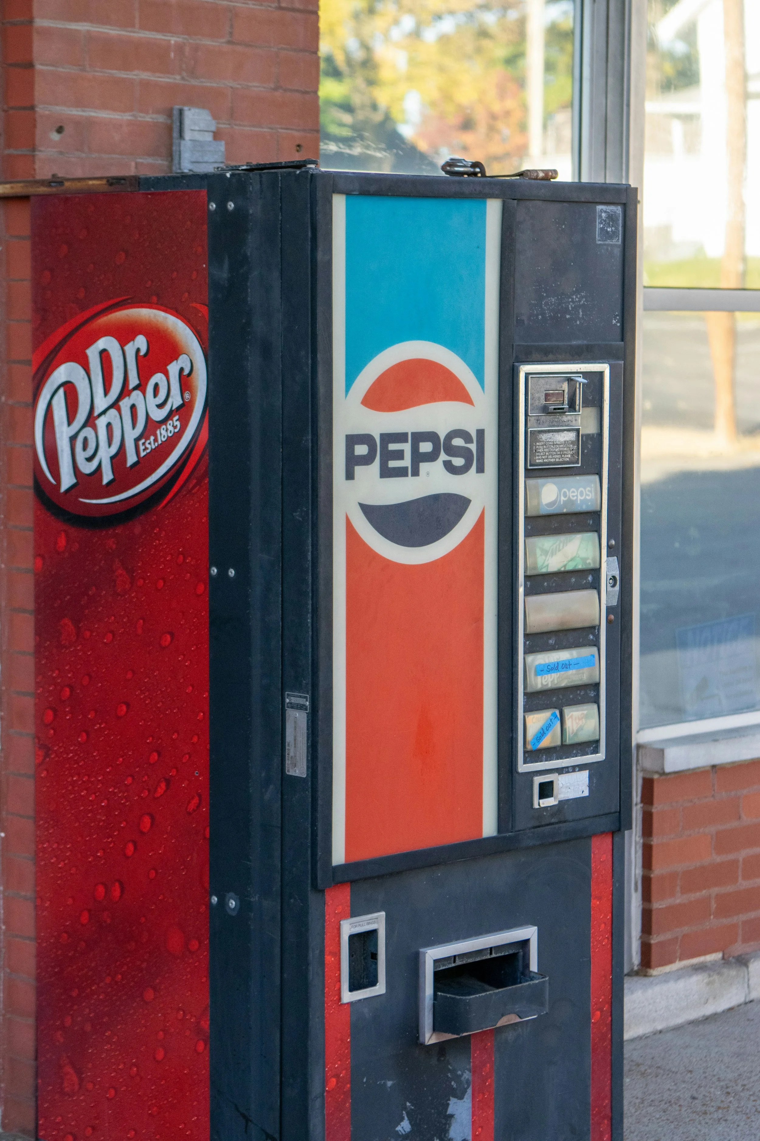 A vending machine with Pepsi branding, located outside a building with a brick wall and window.