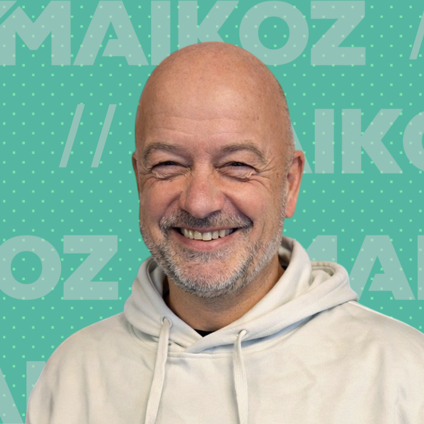 A smiling bald man with a beard wearing a white hoodie, standing in front of a teal background with MAIKOZ text.