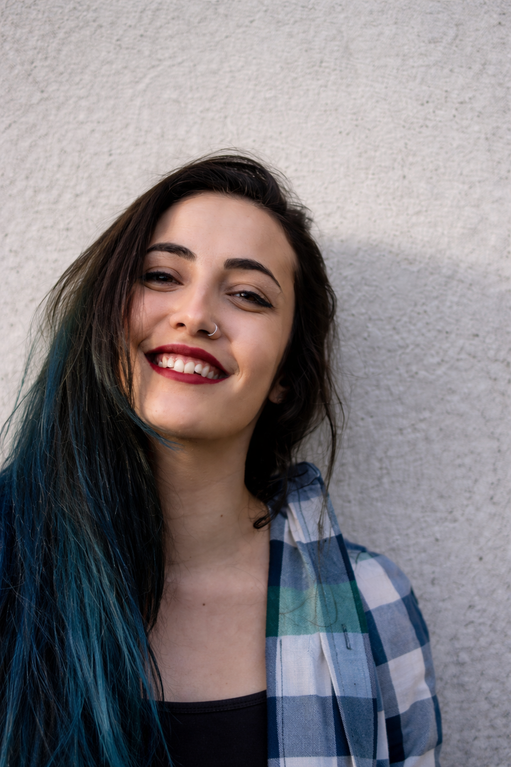 A young woman with dark hair that has blue streaks, smiling and leaning against a textured white wall, wearing a plaid shirt and red lipstick.
