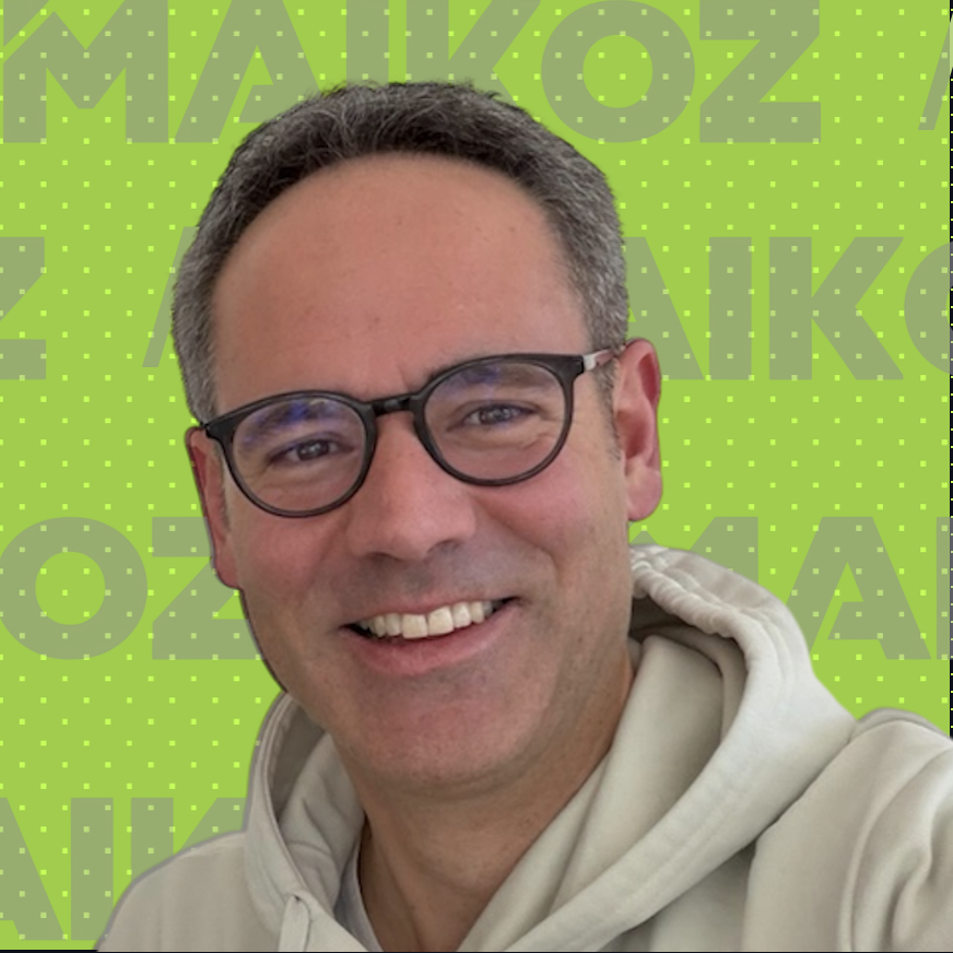 Smiling man with glasses in front of a green background with yellow dots and partially visible MAIKOZ text.