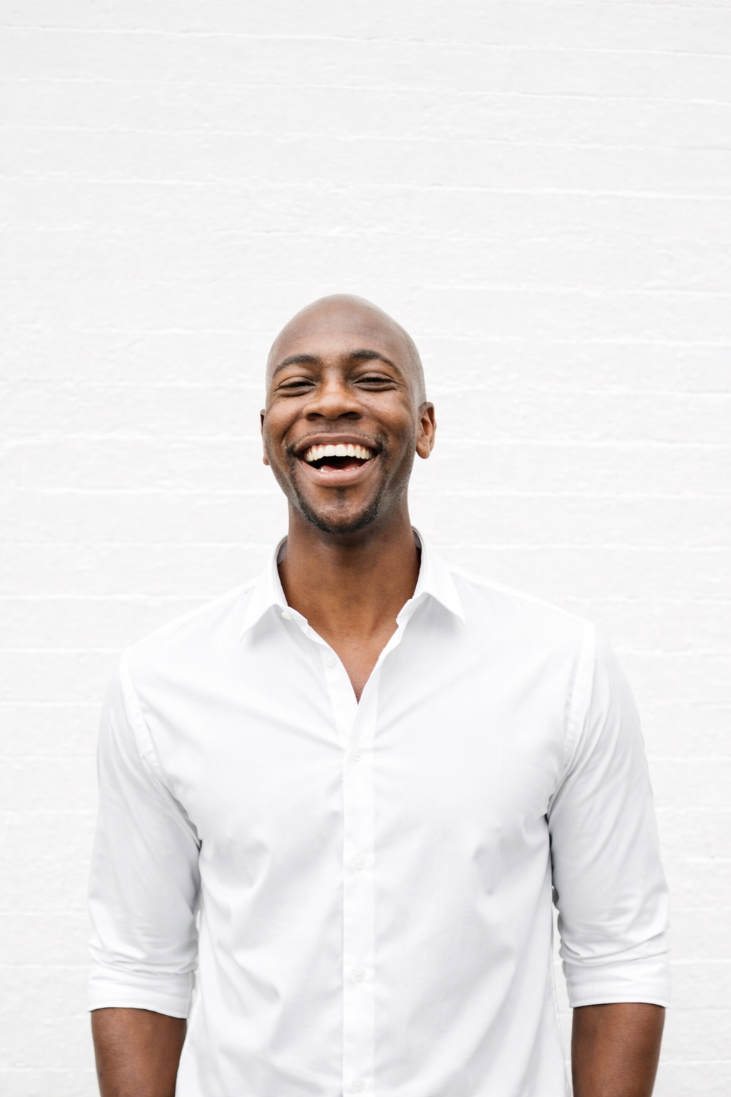 A man with a bald head, wearing a white button-up shirt, smiling and laughing against a white brick wall background.