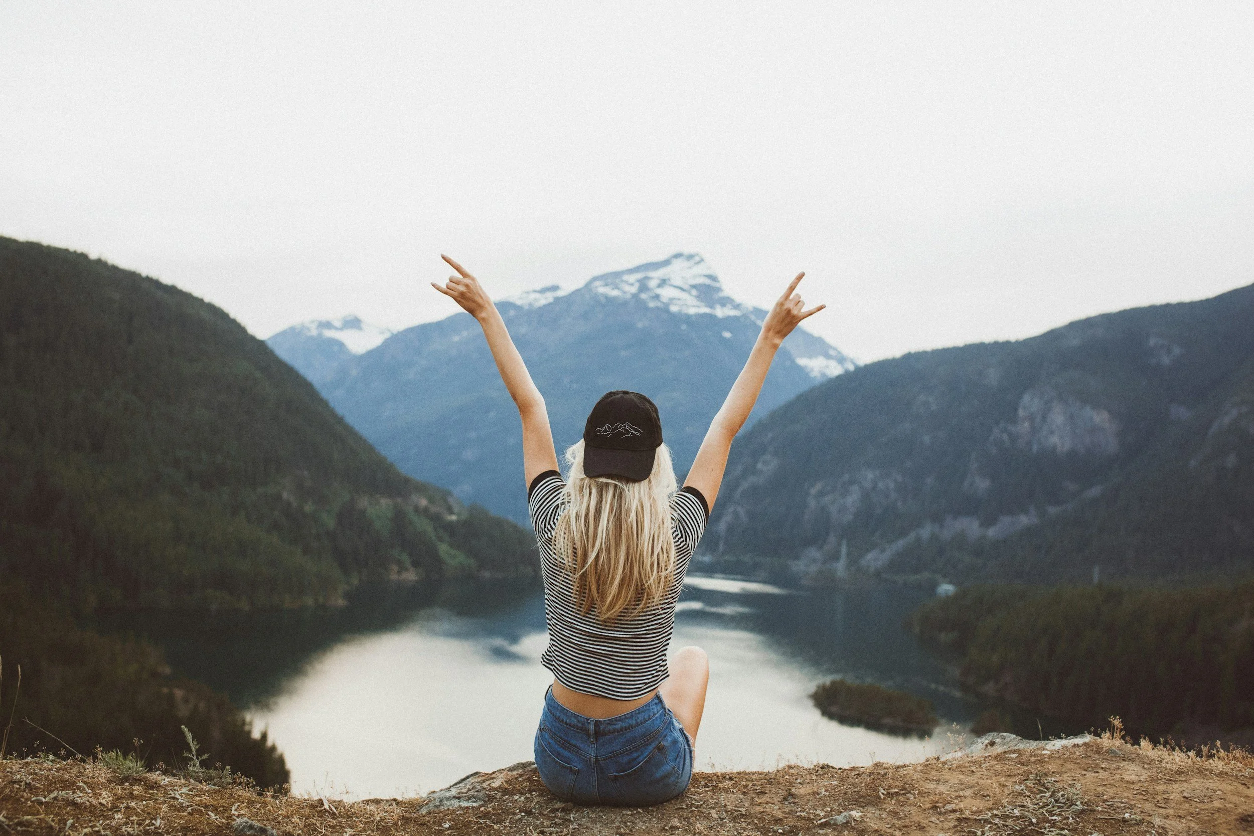 A woman with long blonde hair, wearing a striped shirt, shorts, and a black cap, sits on a rock with her back to the camera, raising her arms in a V shape, overlooking a mountain lake surrounded by forested hills and snow-capped peaks.
