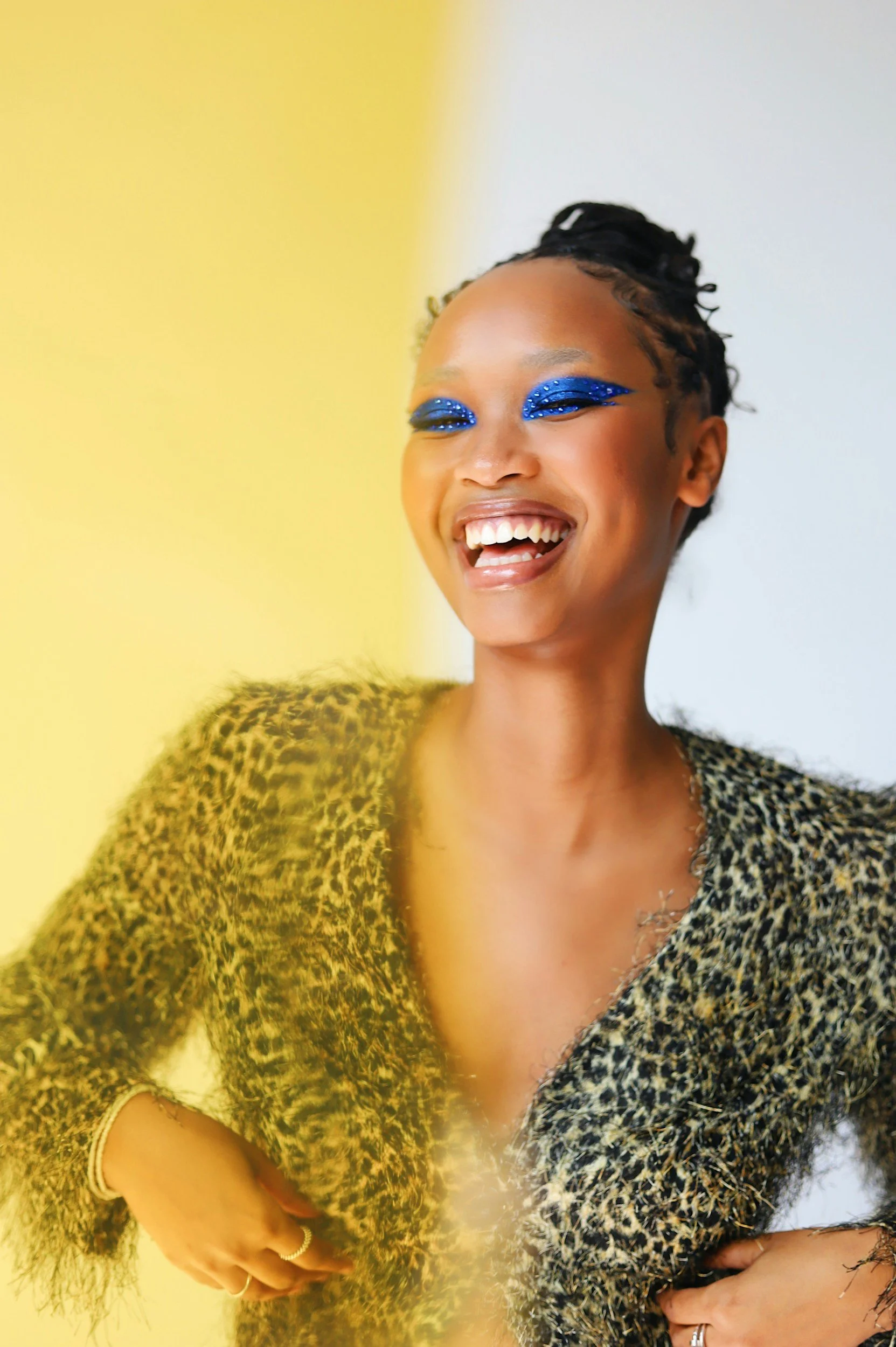 A woman with short dreadlocks and bright blue eye makeup, smiling widely, wearing a leopard print jacket.