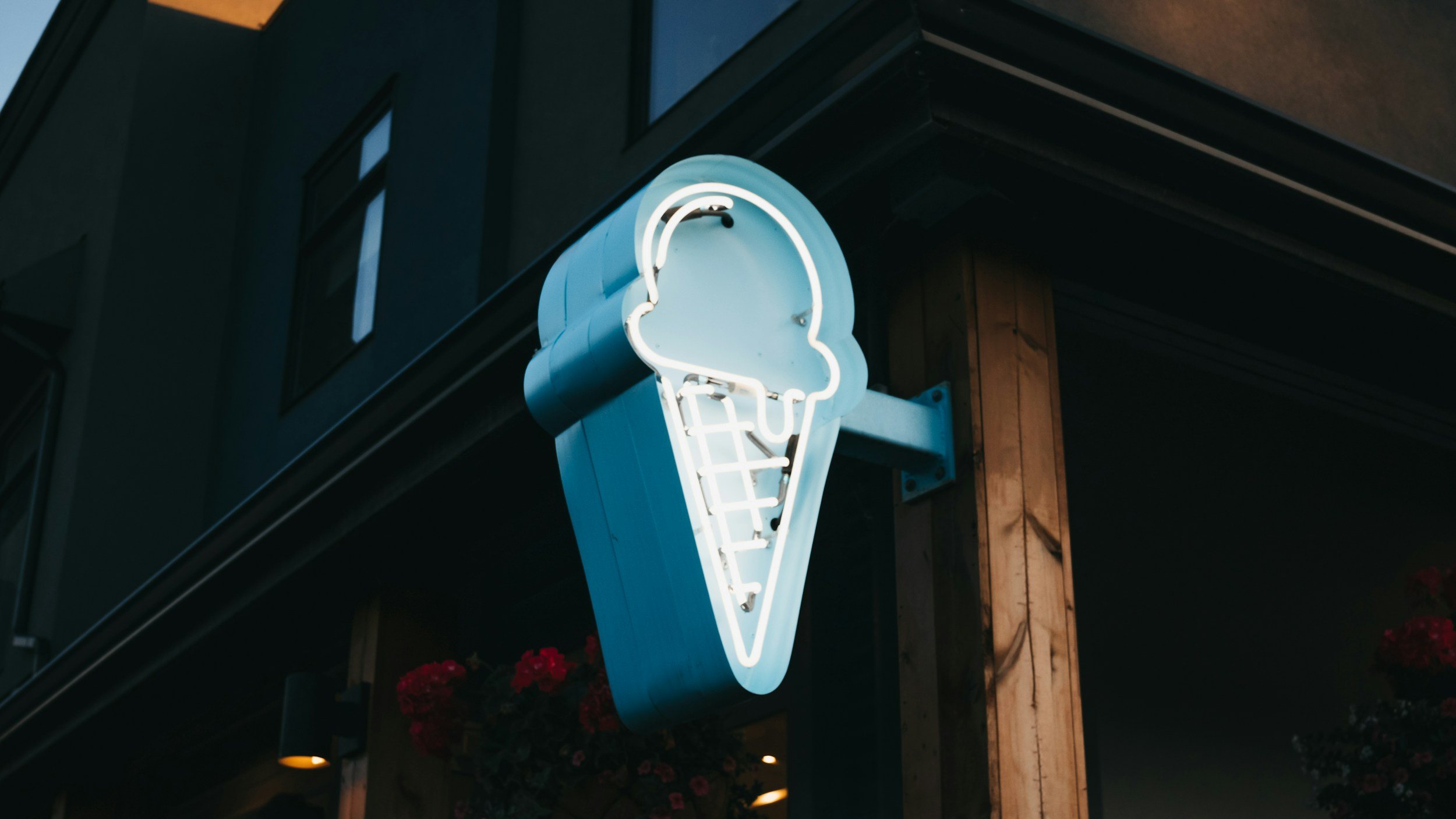 Neon sign in the shape of an ice cream cone mounted on the exterior wall of a building at night, with flowers below.