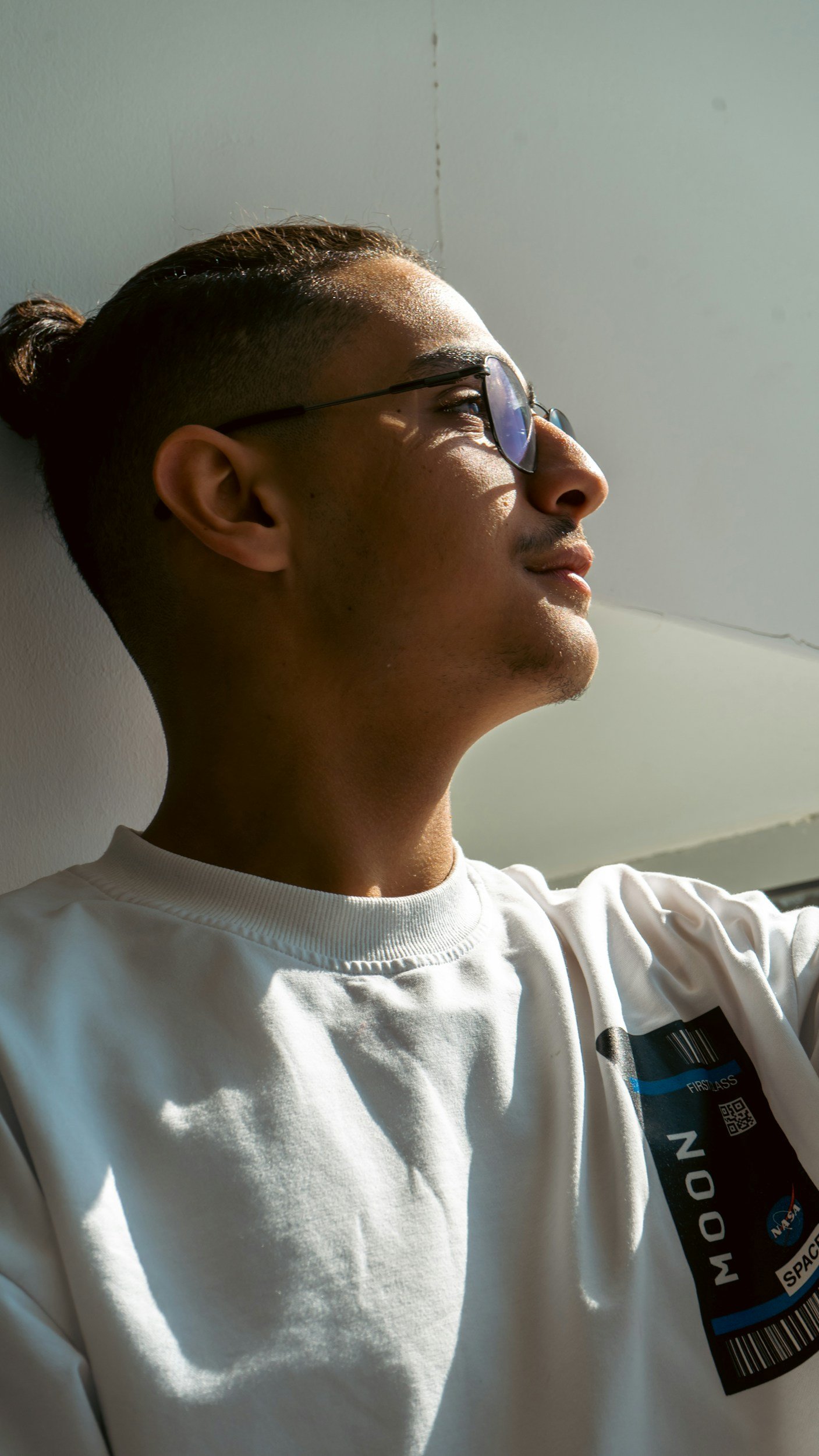A young man with glasses and a slicked-back hairstyle, wearing a white NASA-themed sweatshirt, is sitting against a wall with sunlight illuminating his face.