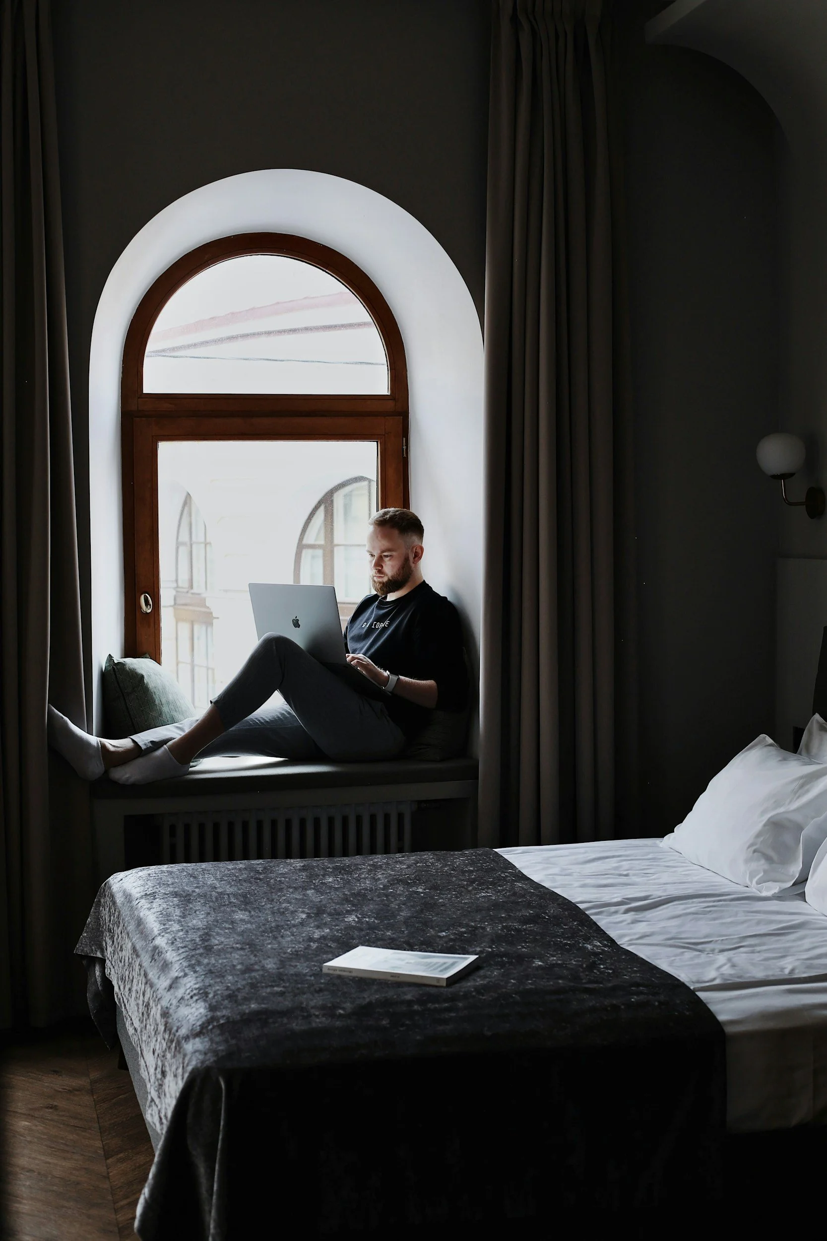 A man with a beard and short hair is sitting on a window seat, working on a laptop. The window behind him has an arched shape with wooden framing, and the room has dark curtains and a lit wall sconce. A bed with a dark velvet blanket and a book on it is in the foreground.