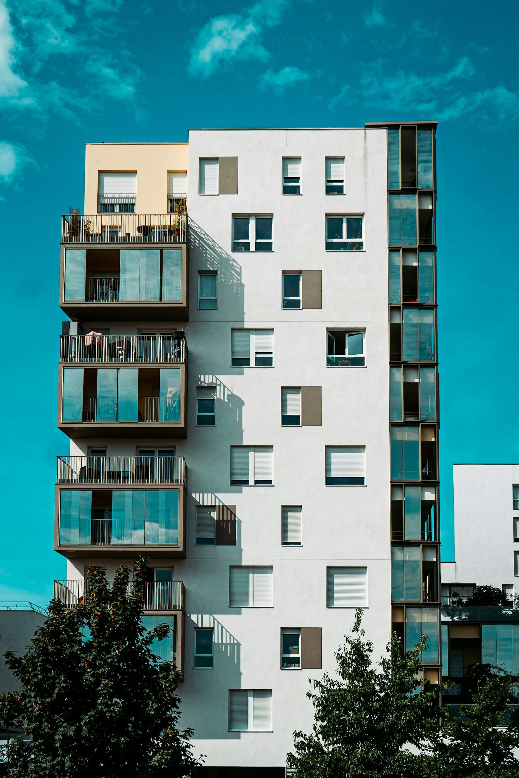 Modern multi-story apartment building with white and beige exterior, featuring balconies with glass railings and variously shaded windows, set against a clear blue sky with a few clouds, and trees at the bottom.