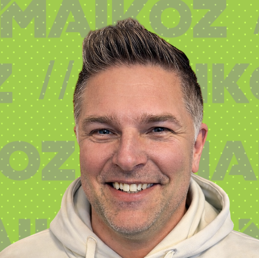 A smiling man with short, styled brown hair wearing a light-colored hoodie, standing in front of a bright green background with MAIKOZ text and dot patterns.