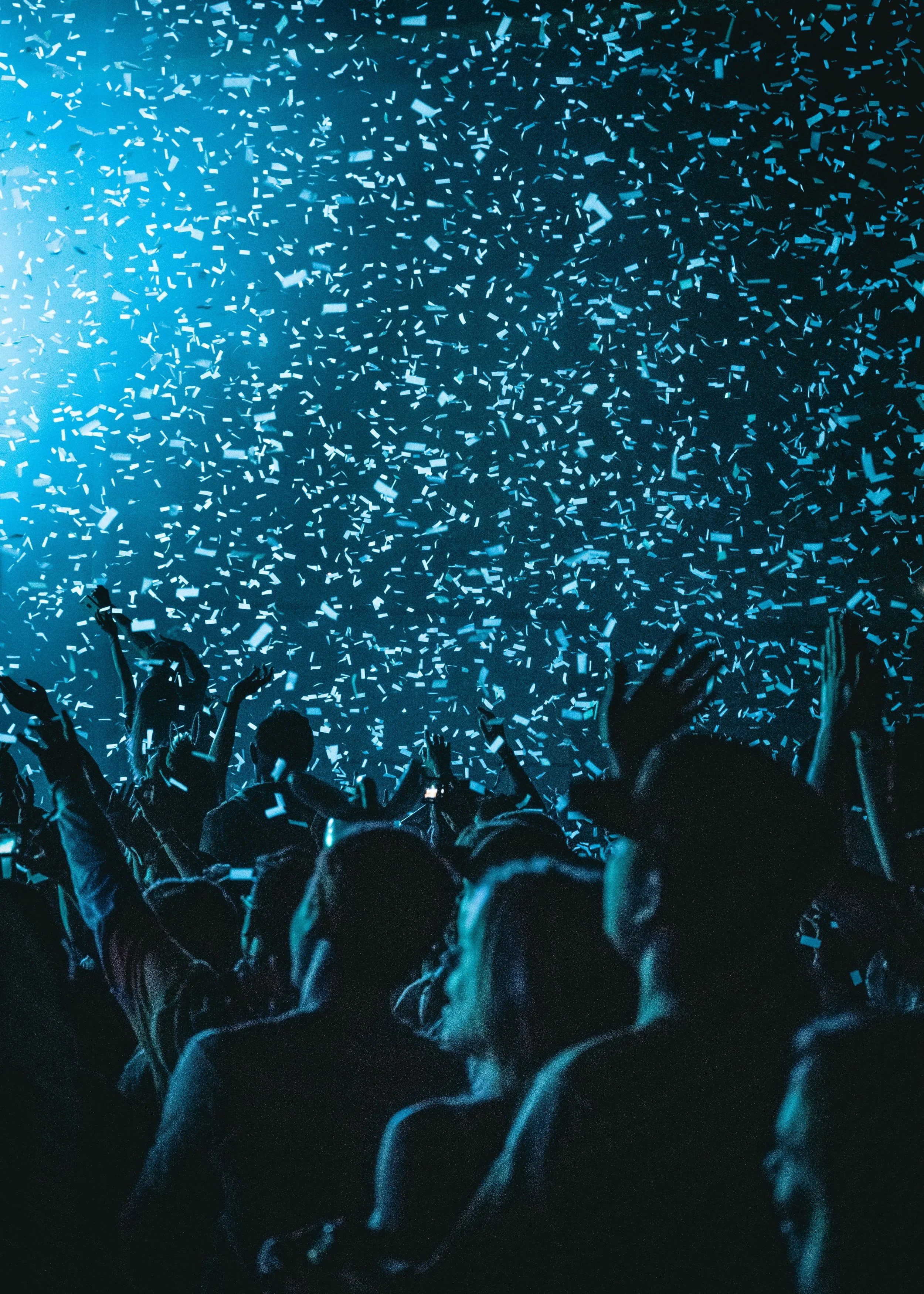 Crowd of people at a concert or event, with confetti falling from above, illuminated by blue lighting.
