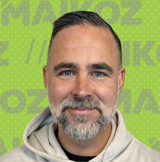 A man with short dark hair, a beard and mustache, smiling, wearing a light-colored hoodie, against a bright green background with large, light green text that says 'MAIKOZ'. 