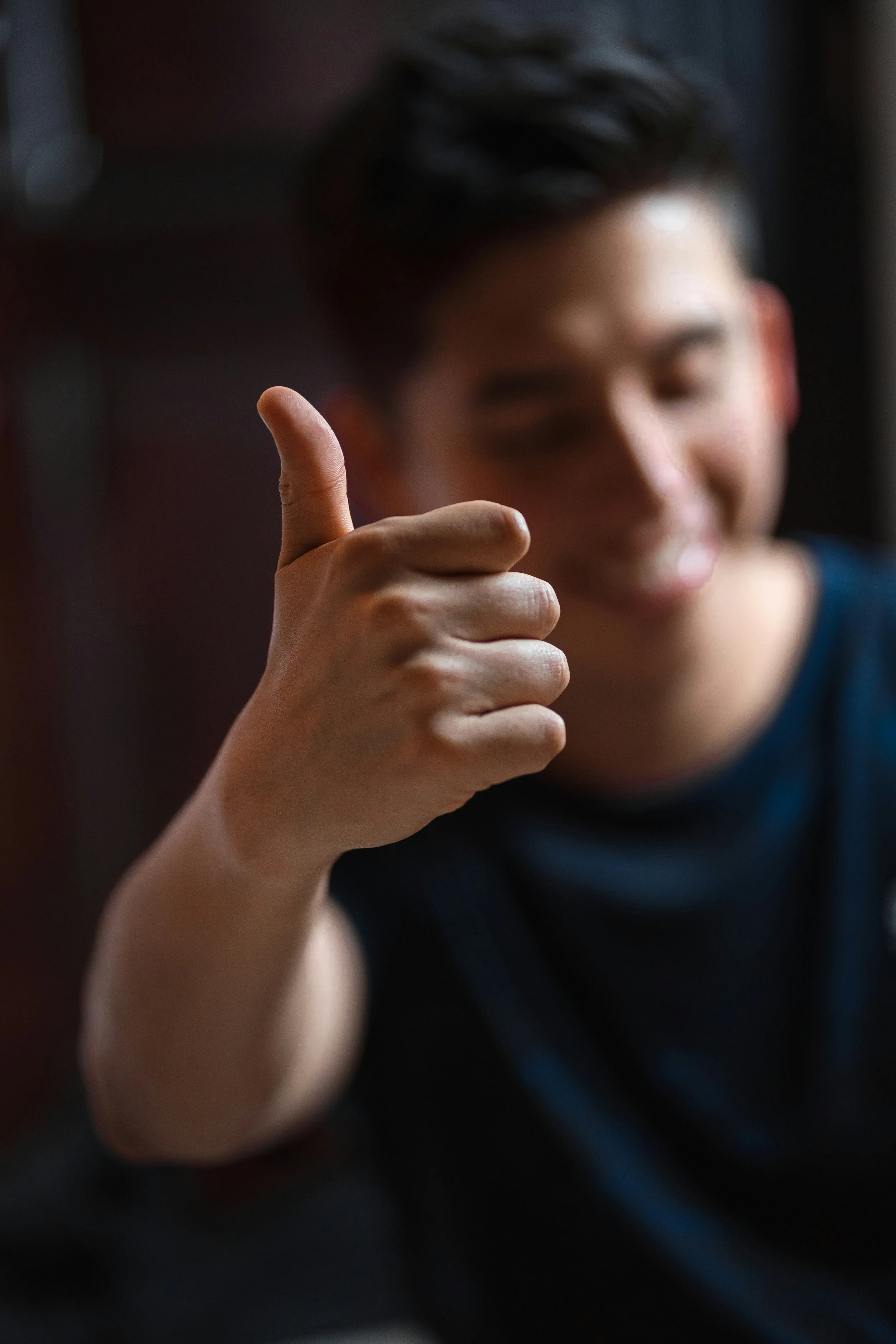 A person giving a thumbs-up gesture, with a blurred smiling face in the background.
