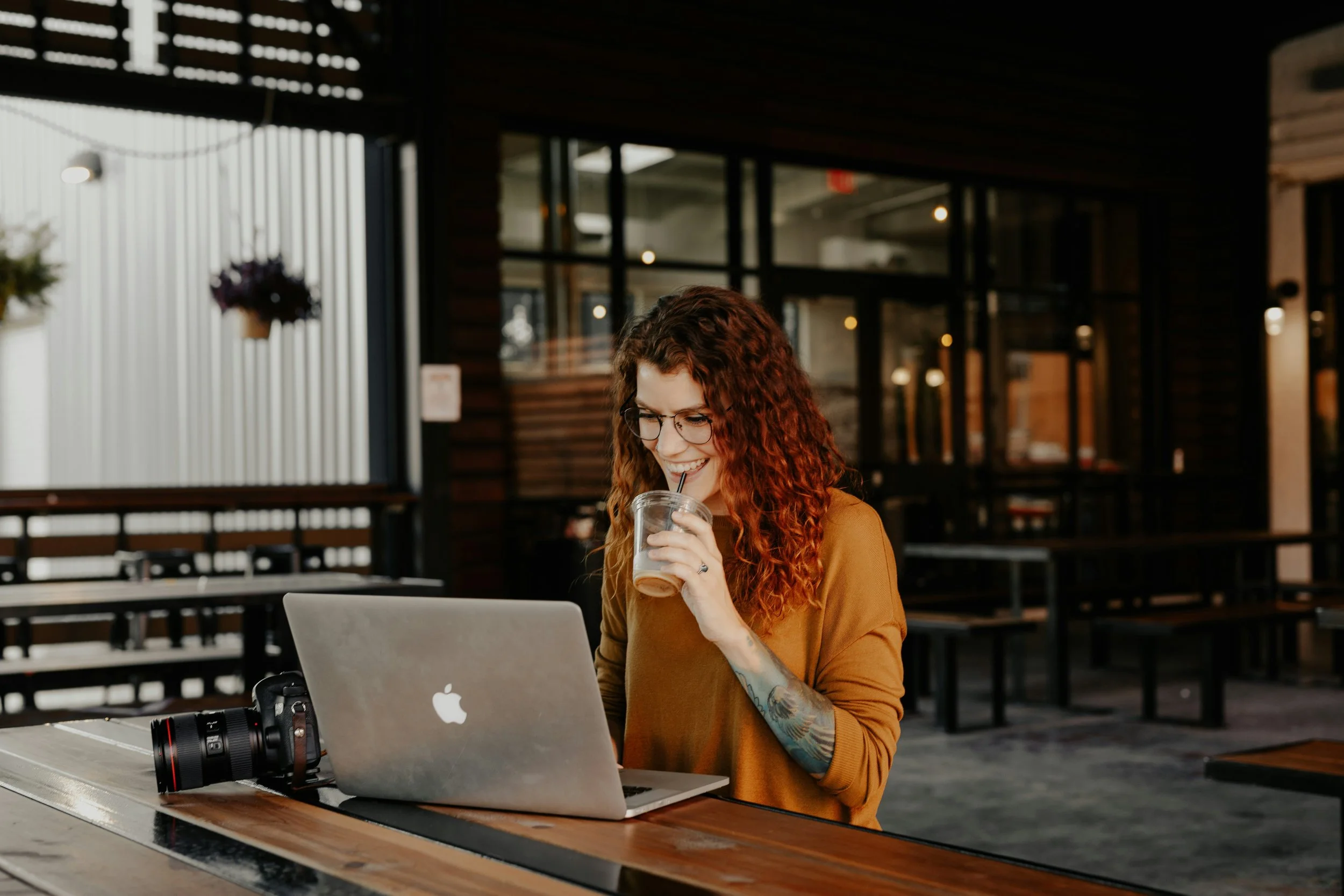 A woman with curly red hair and glasses sitting at a wooden table in a coffee shop, smiling while drinking iced coffee with a straw from a clear glass. A camera is placed next to her laptop.