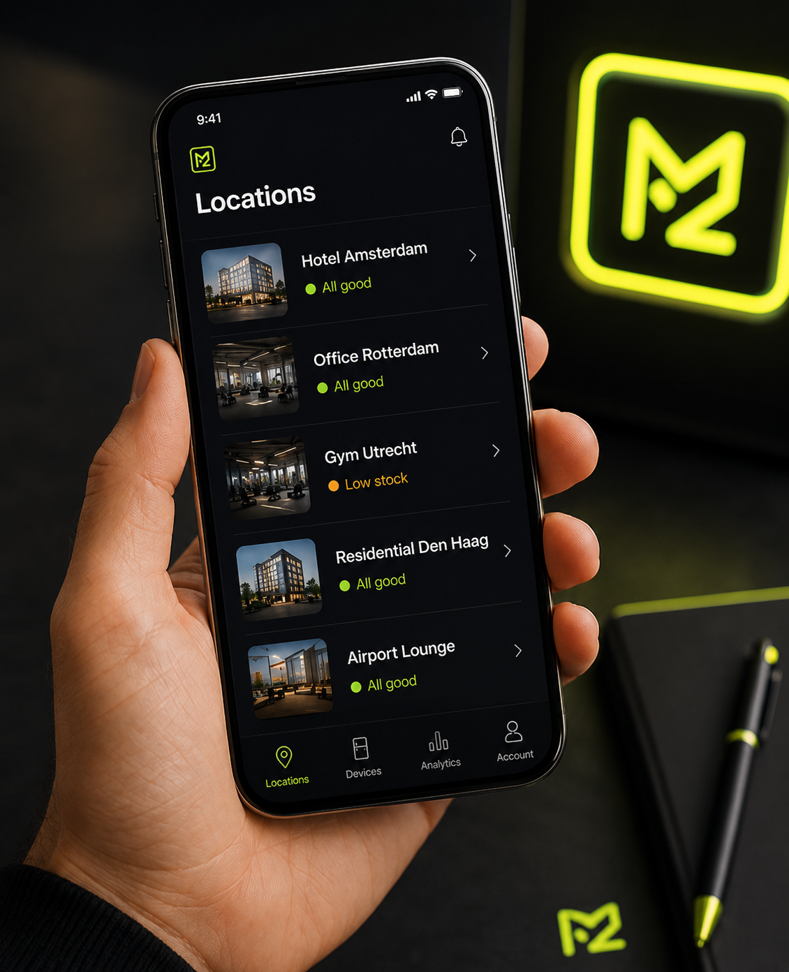 Smartphone screen displaying a location monitoring app with five locations listed, including hotel, office, gym, residential, and airport lounge, with status indicators for each; hand holding phone; dark background with sign and branding items visible.