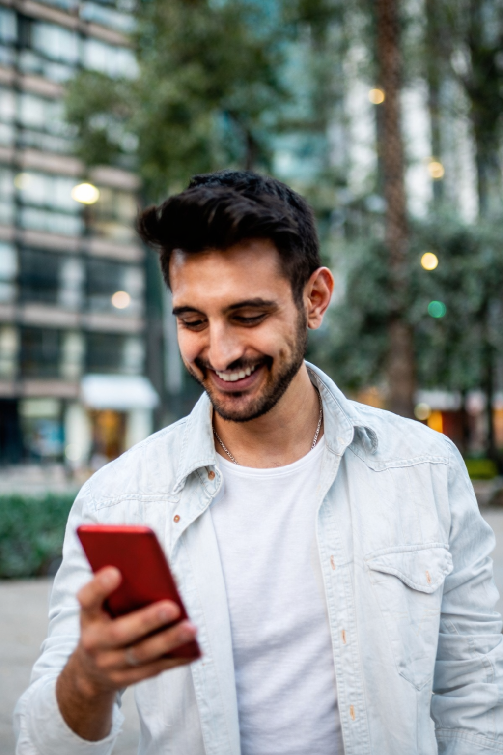 A smiling young man with dark hair, beard, and mustache, wearing a light denim shirt over a white T-shirt, looking at his red smartphone outdoors in an urban park setting with trees, buildings, and blurred city lights in the background.