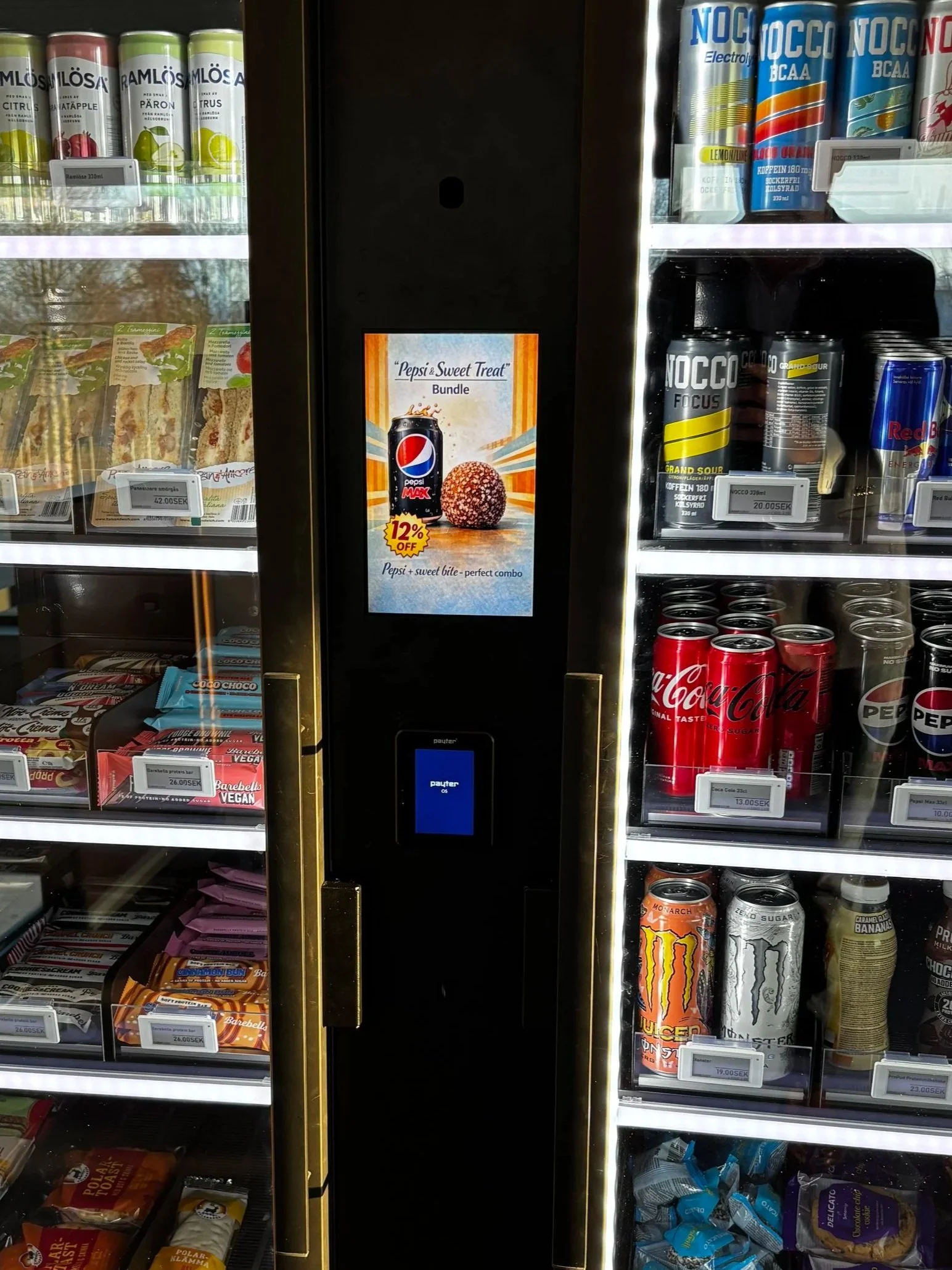 Vending machine stocked with snacks and beverages, including energy drinks, soda cans, candy bars, and protein drinks.