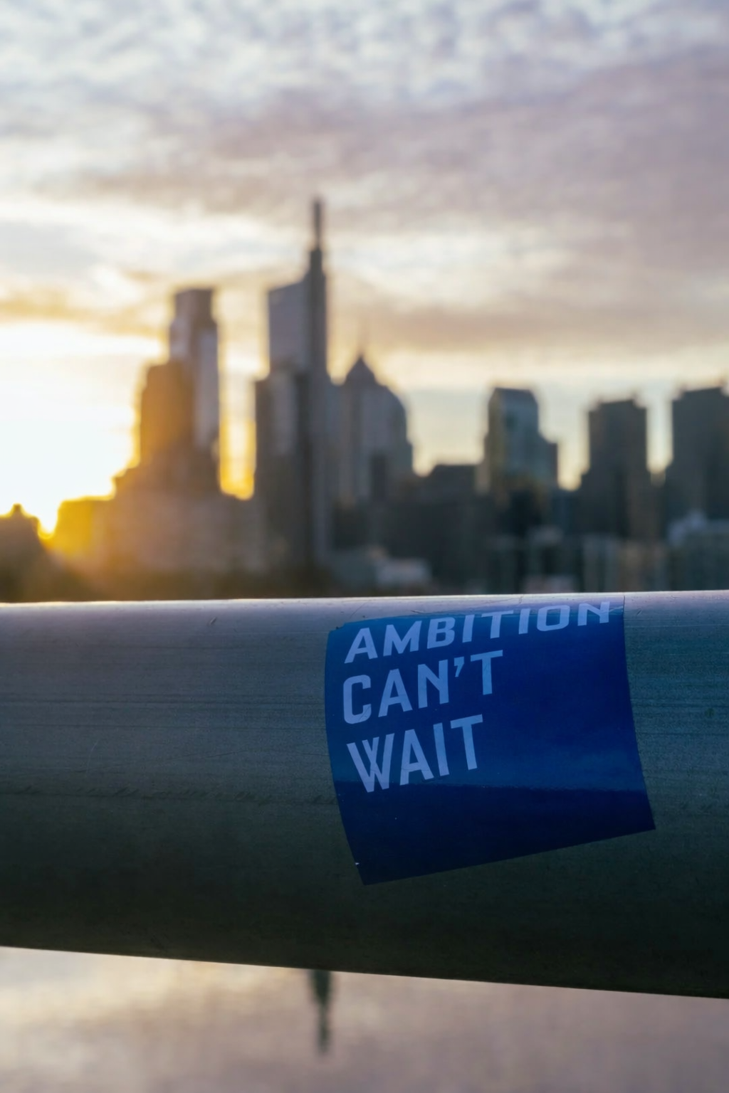 A city skyline blurred in the background during sunset or sunrise, with a focus on a railing in the foreground that has a blue sticker reading 'AMBITION CAN'T WAIT'.