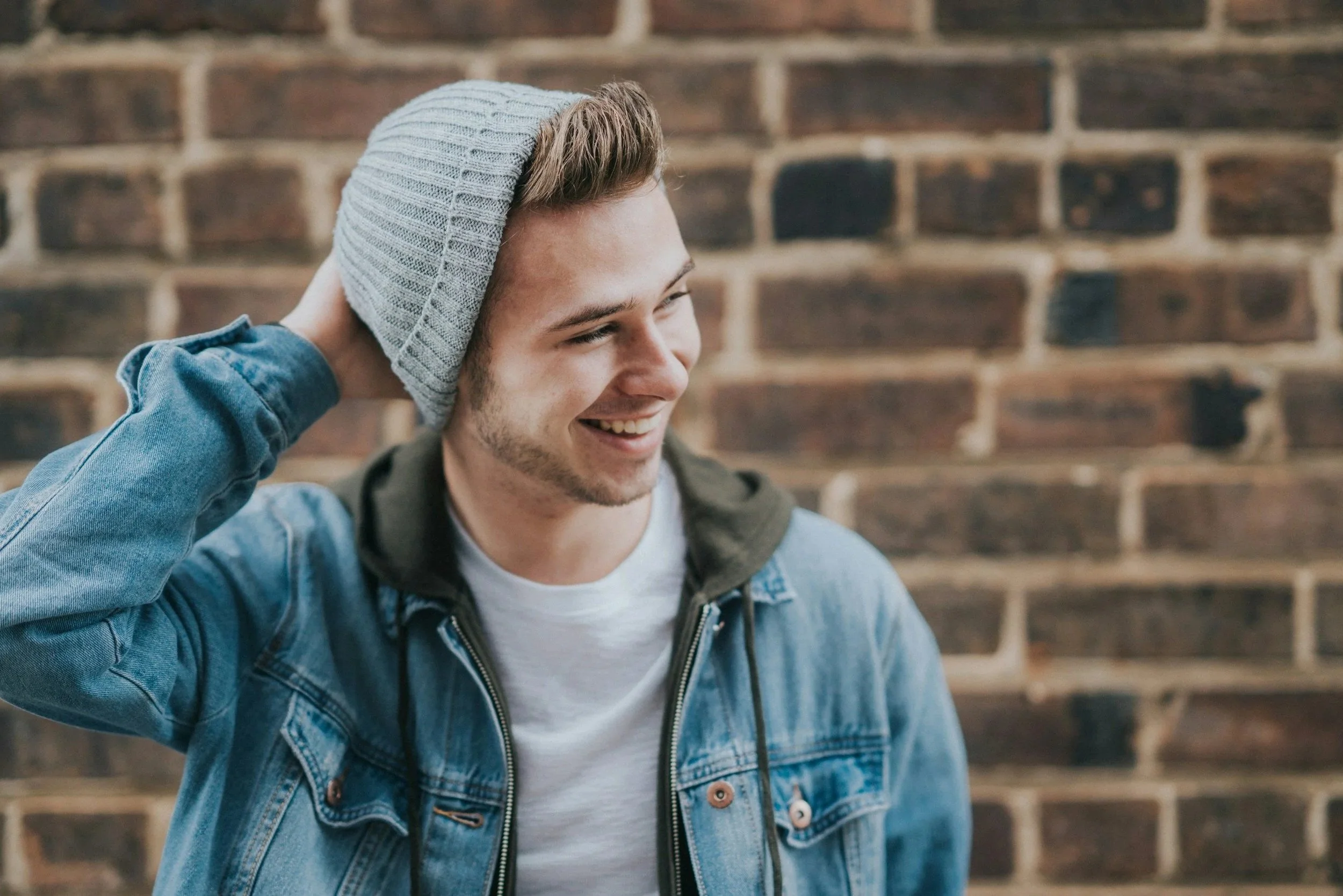 A young man with a light beard and mustache smiling, wearing a gray knit beanie, a denim jacket over a hoodie, and a white t-shirt, standing against a brick wall.