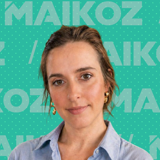 A woman with shoulder-length brown hair wearing a light blue shirt and gold hoop earrings, standing in front of a turquoise background with the text 'MAIKOZ' repeatedly printed in large, light-colored letters.