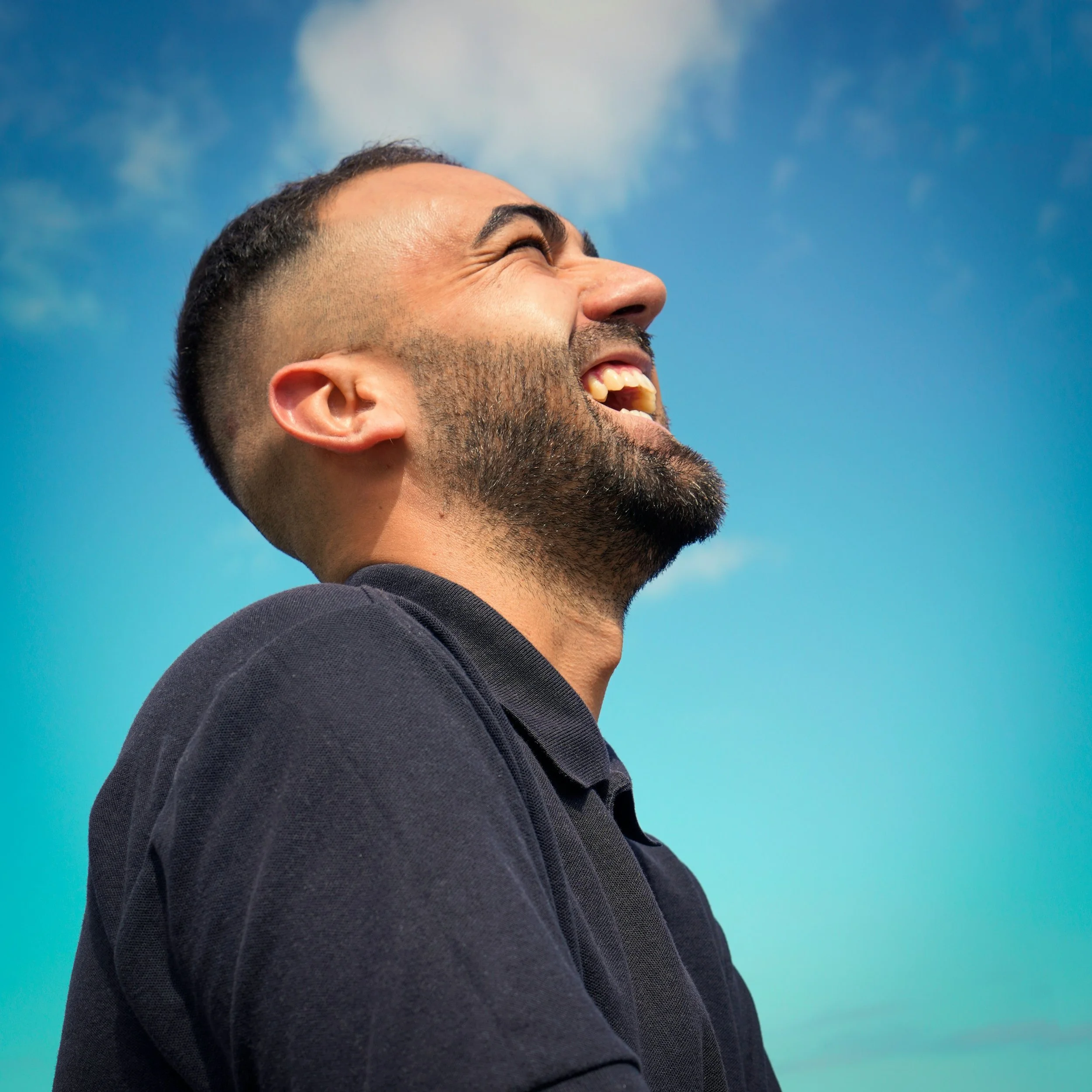 Close-up of a man smiling and looking up against a blue sky with some clouds.