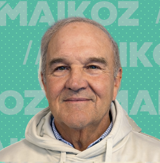 A middle-aged man with gray hair and blue eyes, wearing a light-colored hoodie and standing in front of a teal background with the word 'MAIKOZ' written repeatedly.
