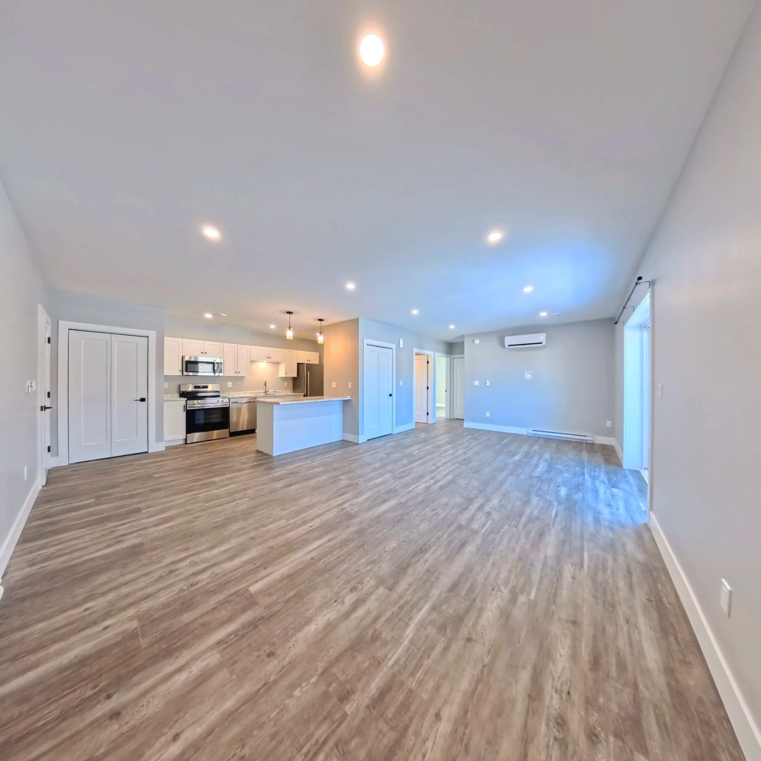A spacious open-concept kitchen and living area with light-colored walls and ceiling, hardwood flooring, and recessed lighting. The kitchen features white cabinets, stainless steel appliances, and a central island. There are two pendant lights hanging above the island. Large windows allow natural light into the room.