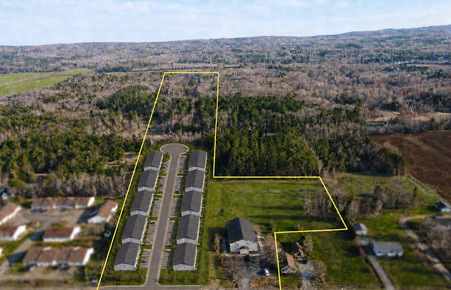 Aerial view of a residential area with a row of detached townhouses on a street, adjacent to a large green lawn and surrounded by a densely wooded area, with additional houses and open land in the background.