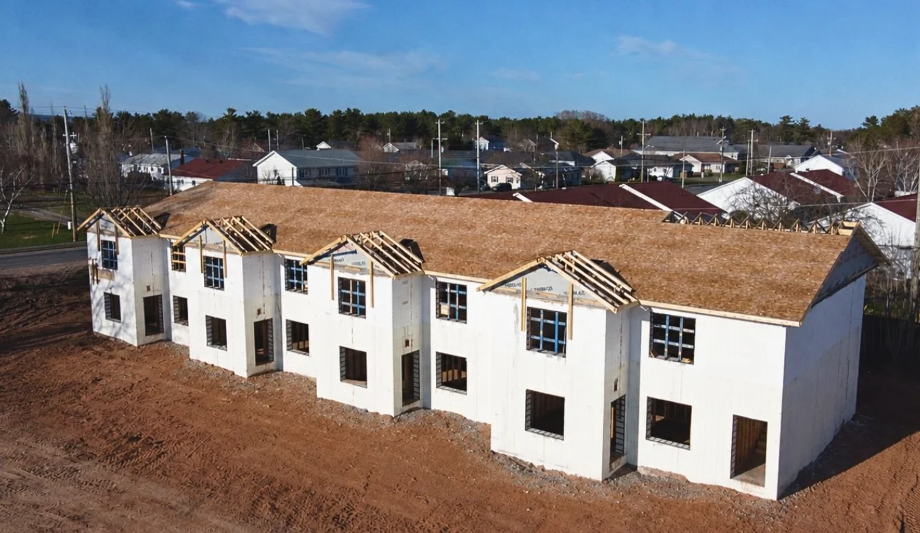 A multi-unit residential building under construction with exterior walls and roof framing in place, but windows, doors, and finishing not yet installed.
