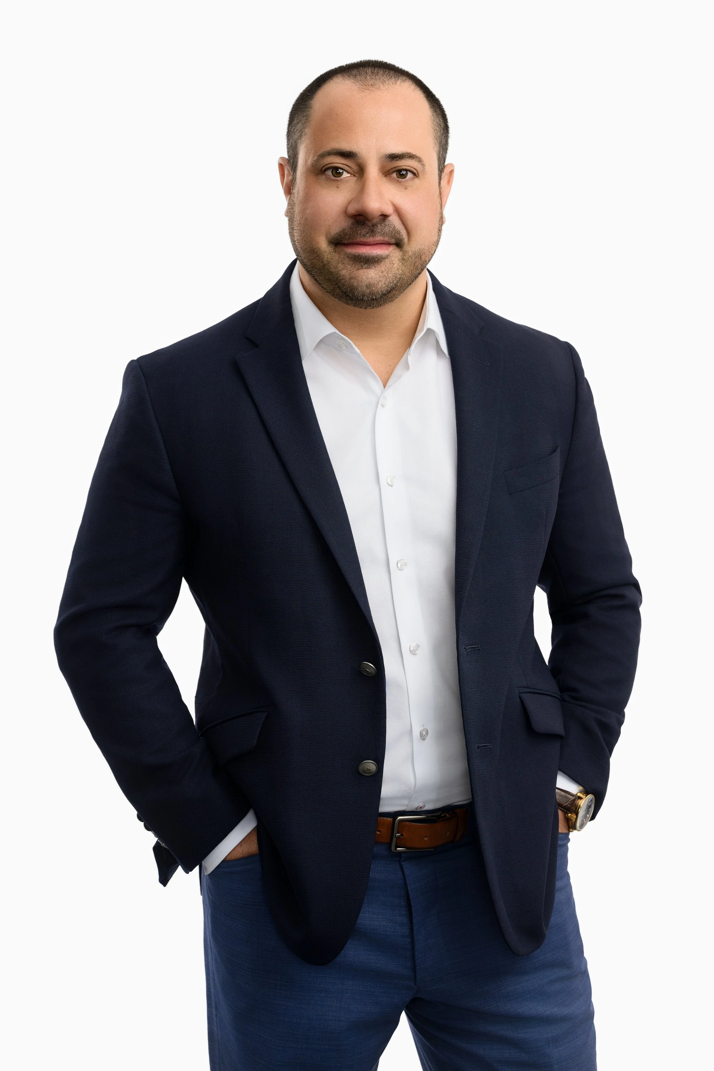 A man with short dark hair, a beard, wearing a dark blue blazer, white shirt, and blue pants, standing against a plain white background.