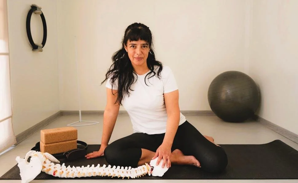 Woman sitting on yoga mat in a fitness or therapy room with exercise equipment, including a stability ball, yoga blocks, and a massage or therapy tool.