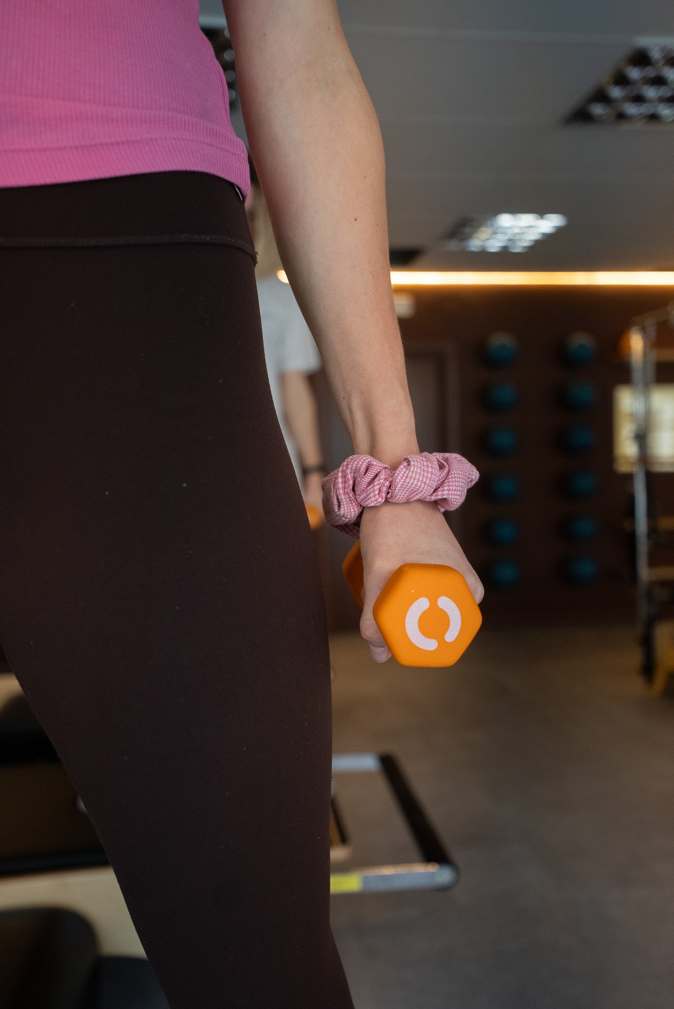 A person wearing a pink scrunchie on their wrist is holding an orange dumbbell in a gym, with gym equipment visible in the background.