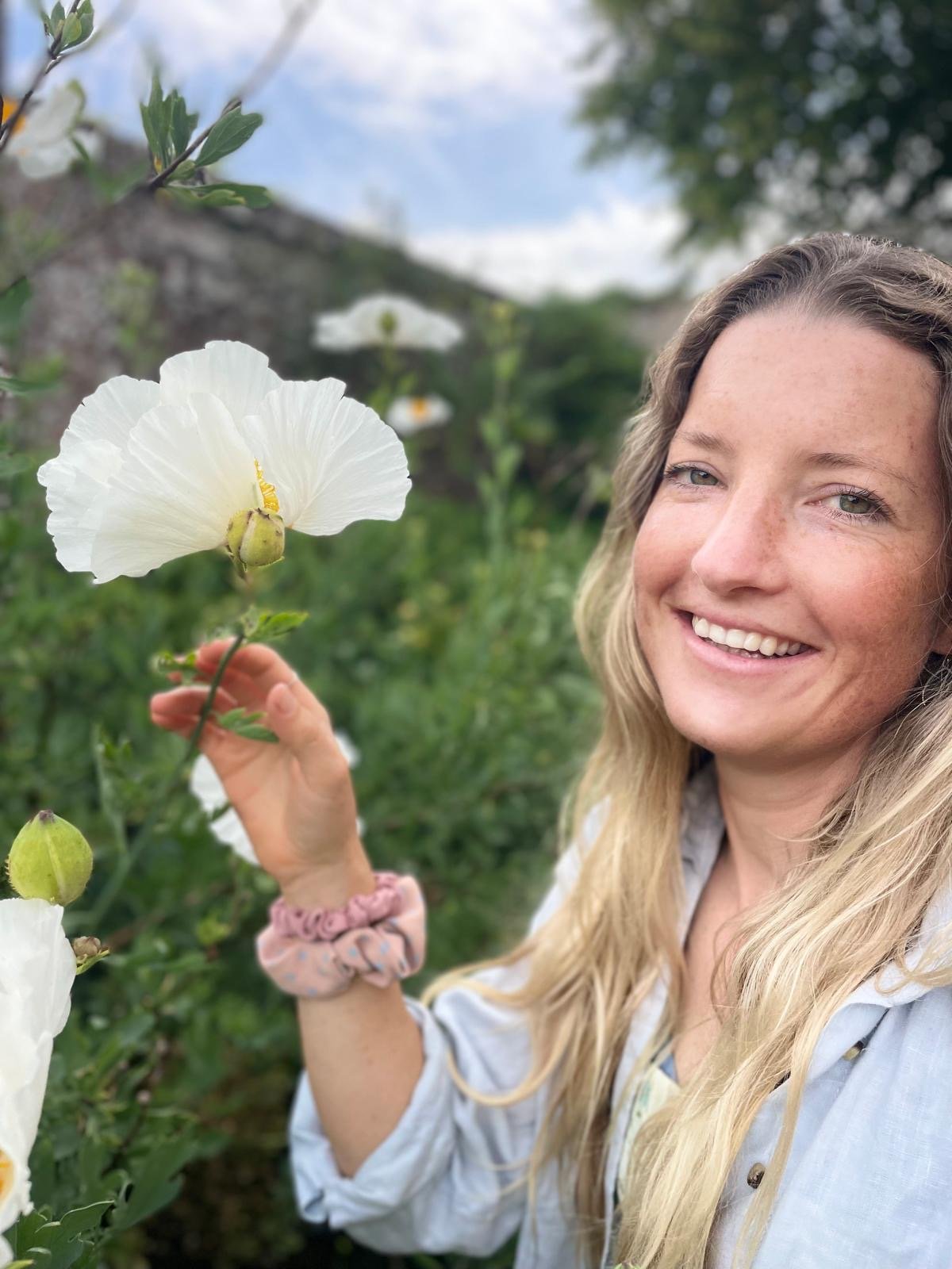 Woman smiling outdoors holding a white hibiscus flower in a garden