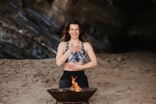 Woman standing outdoors near a rock formation, smiling while holding a stick over a flame in a fire pit.