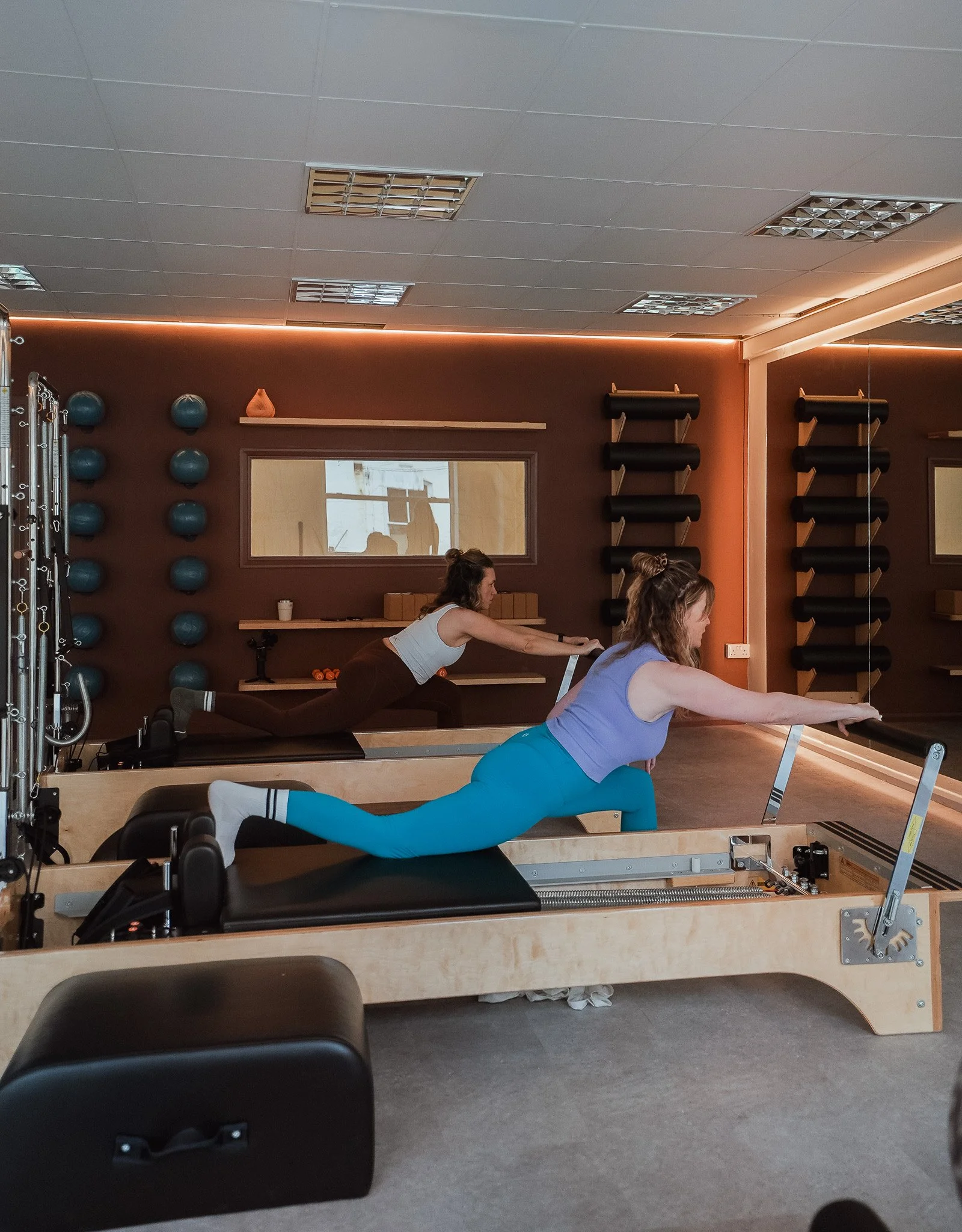 Two women practicing Pilates on reformer machines in a fitness studio with mirrors and wall-mounted exercise equipment.