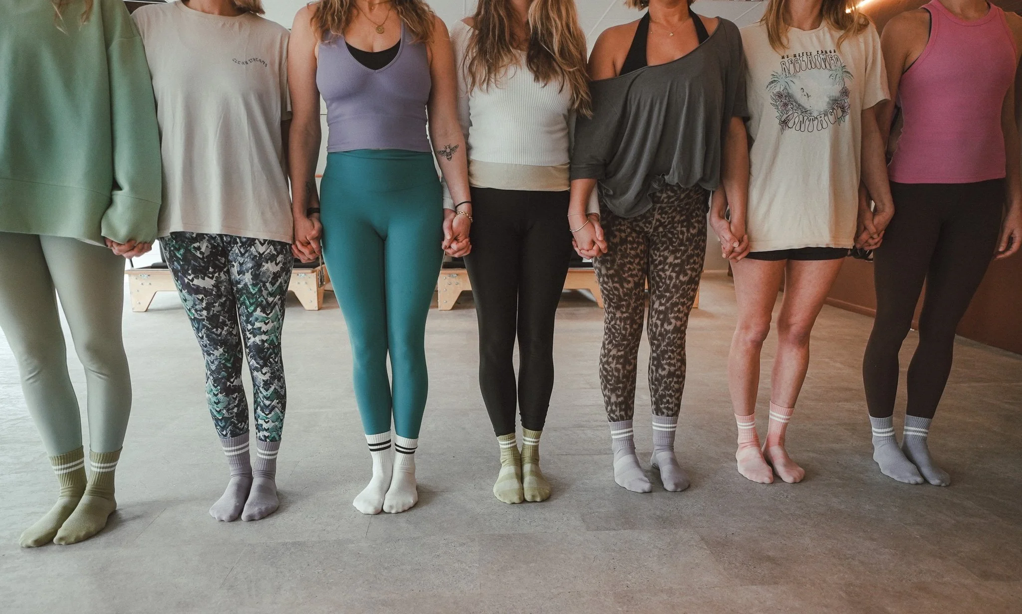 Six women standing in a line holding hands in a yoga or dance studio, wearing workout clothes and socks.