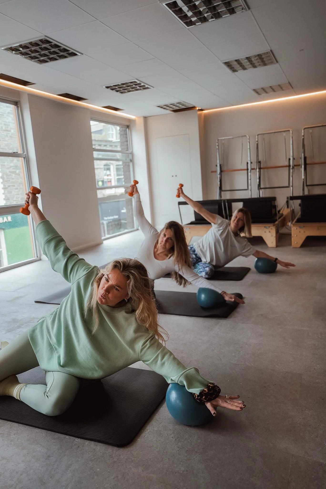 Three women are doing a yoga or exercise class using small dumbbells and exercise balls in a bright, spacious room with large windows, on black mats.