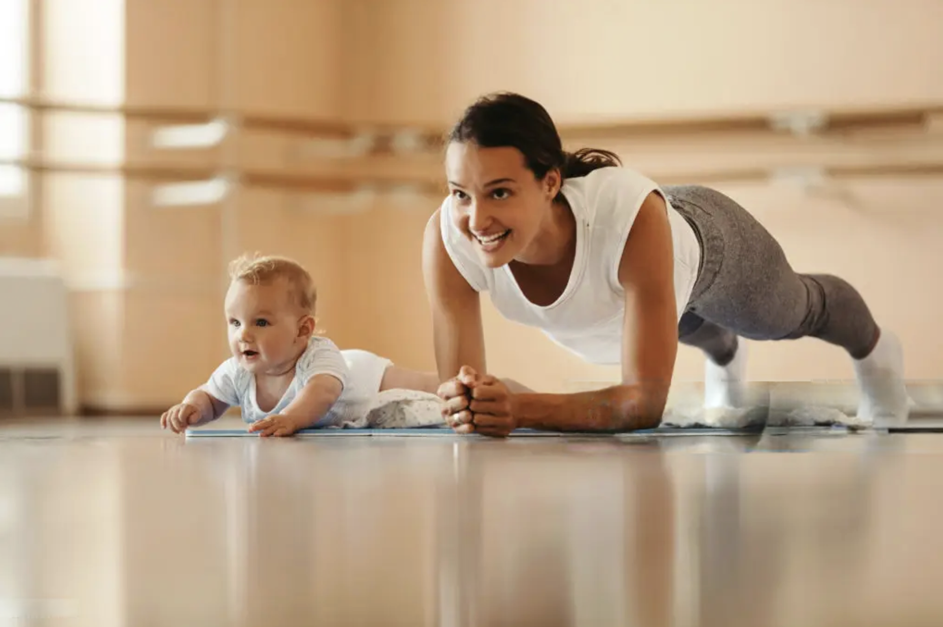 A woman and a baby practicing yoga on the floor in a bright room, both in plank poses, smiling and engaging in the activity.