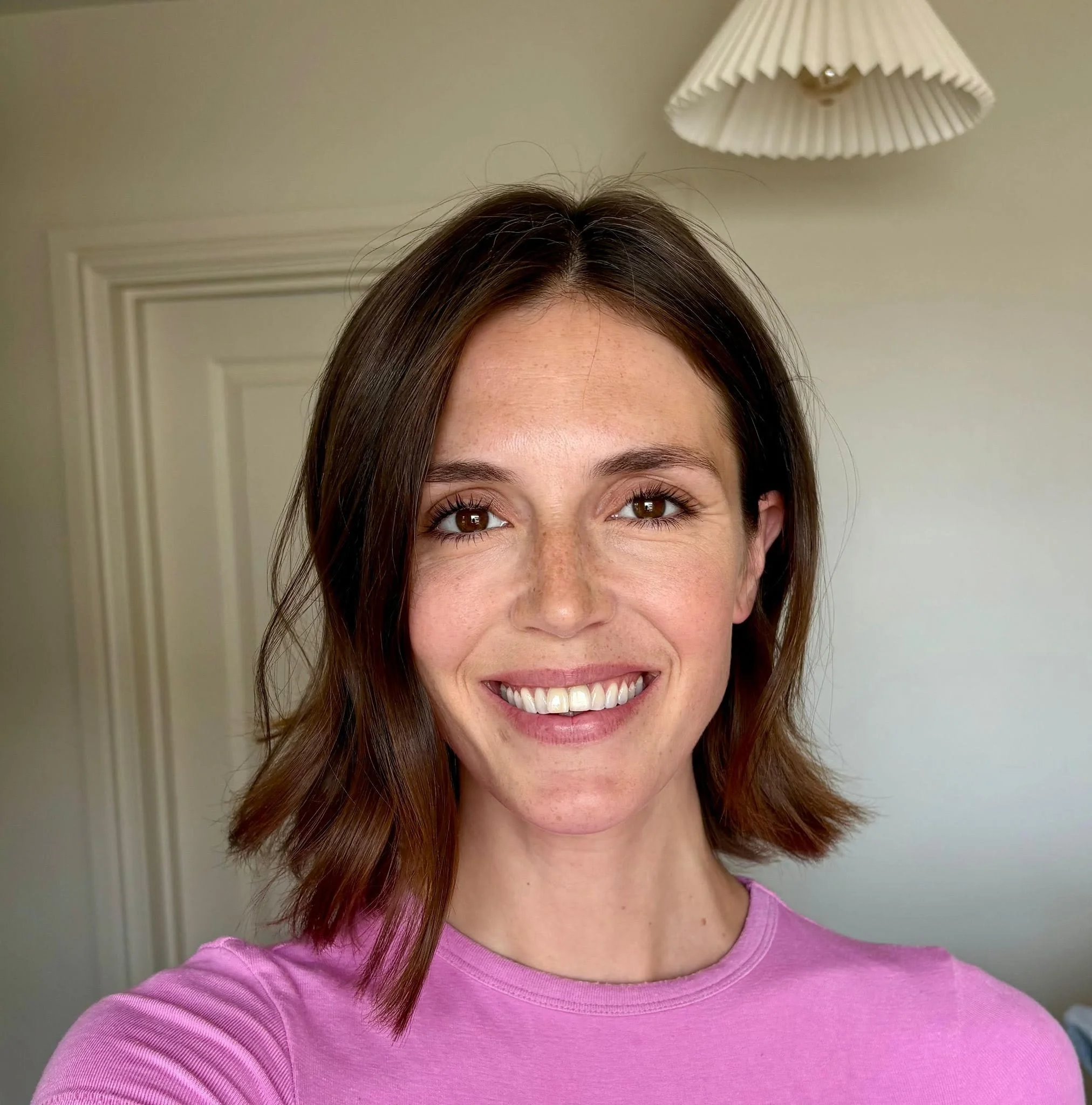 A woman with brown hair, brown eyes, and a broad smile wearing a pink top. She is indoors with a cream-colored wall, a door, and a hanging lampshade in the background.