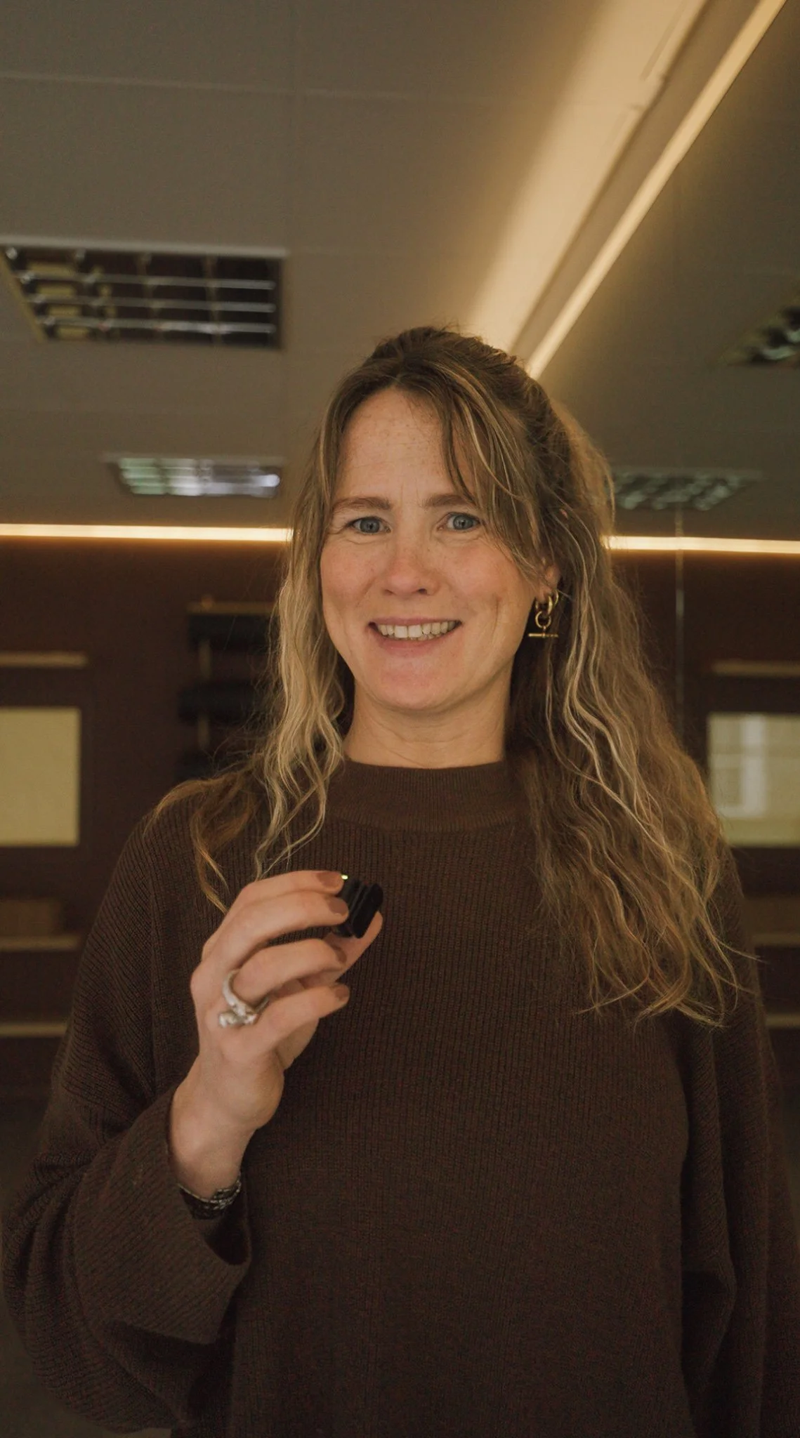 A woman with long wavy hair smiling, wearing a dark brown sweater and gold hoop earrings, standing in an office.
