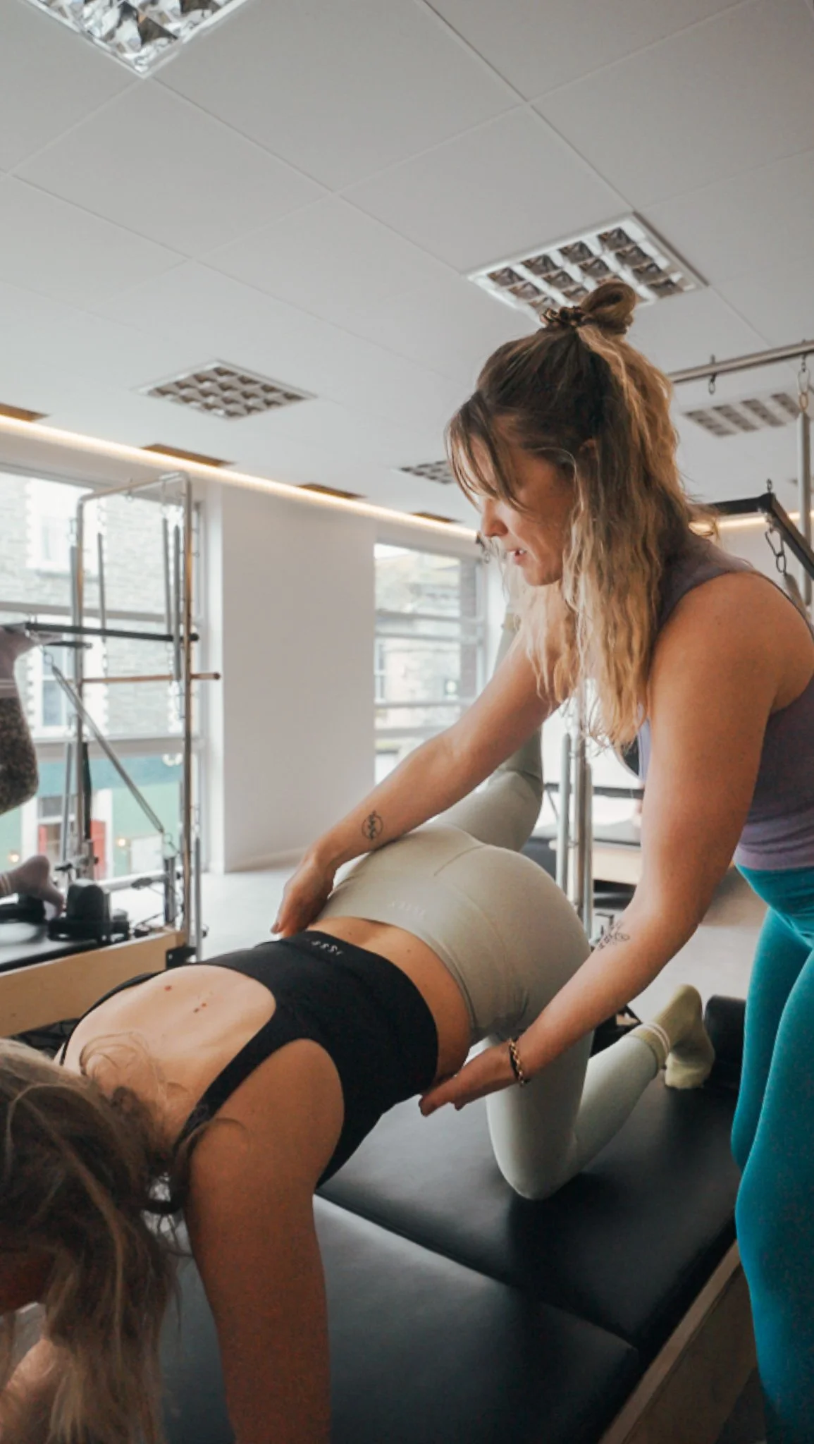 A woman assisting another woman in a yoga pose in a fitness studio with large windows and gym equipment visible.