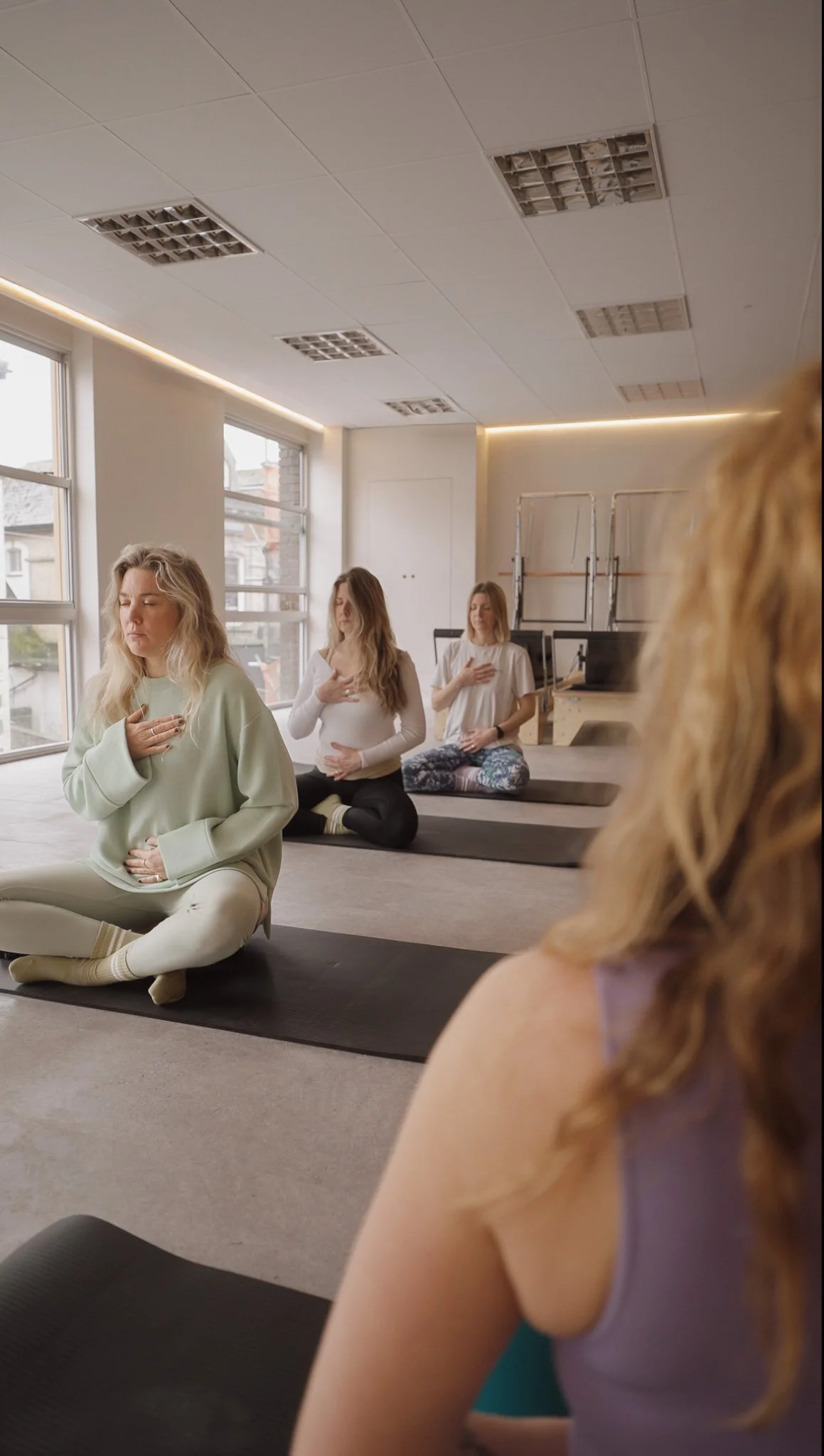 Women practicing meditation in a yoga studio, seated on yoga mats in a cross-legged position with hands on their chests and hearts.