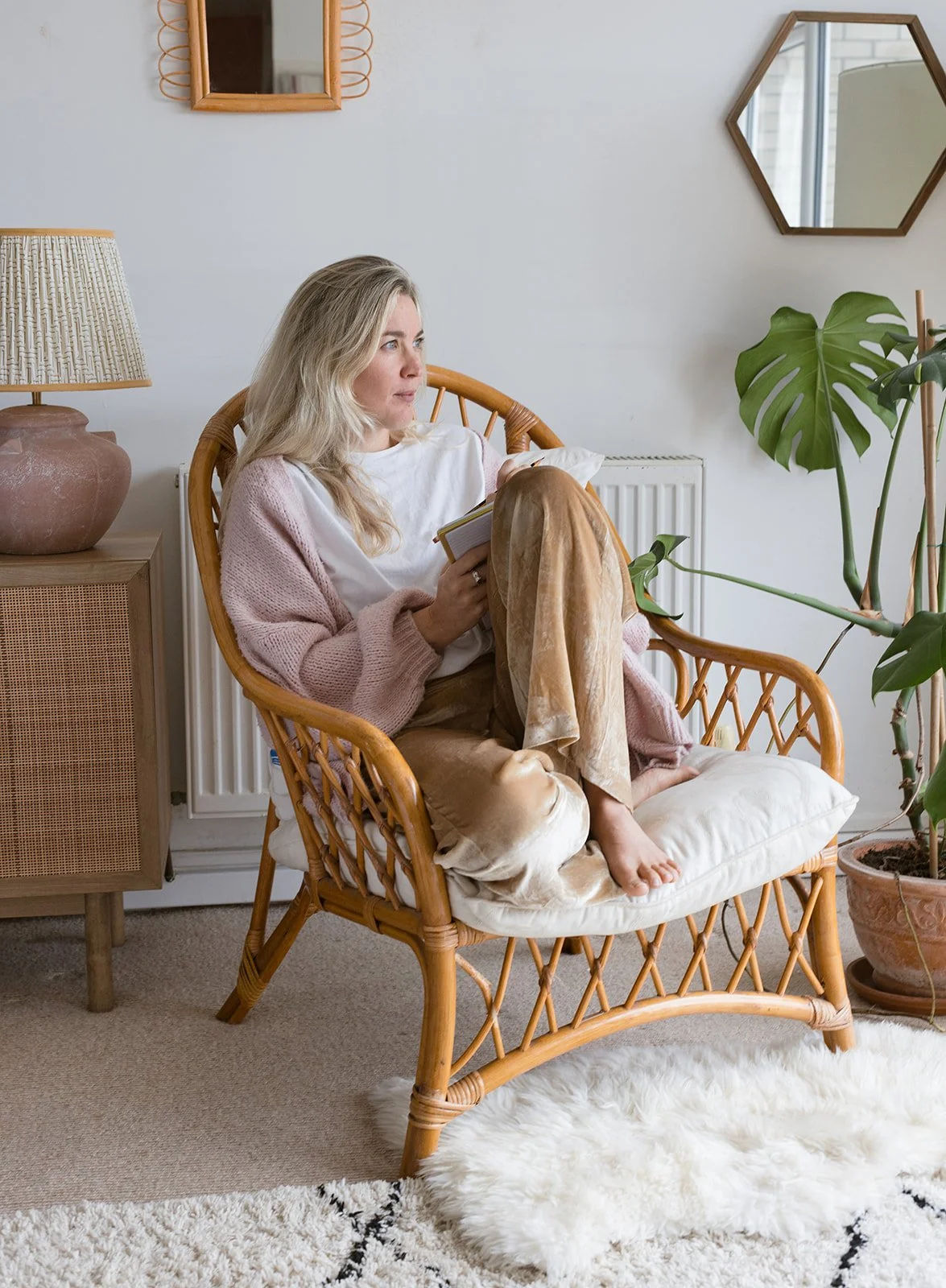 A woman with long blonde hair sitting on a wicker chair, reading a notebook, in a cozy living room decorated with indoor plants, a table lamp, a wall mirror, and a white fluffy rug.