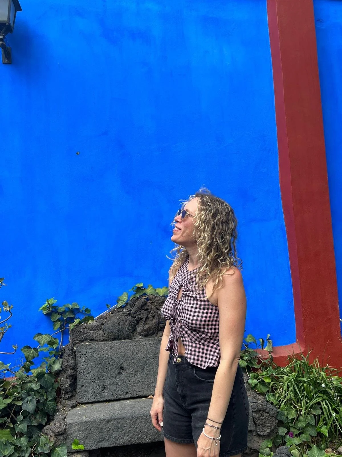 Woman with curly blonde hair and sunglasses smiling, wearing a sleeveless gingham top tied in front and black shorts, standing outdoors against a bright blue wall with plants and rocks around.