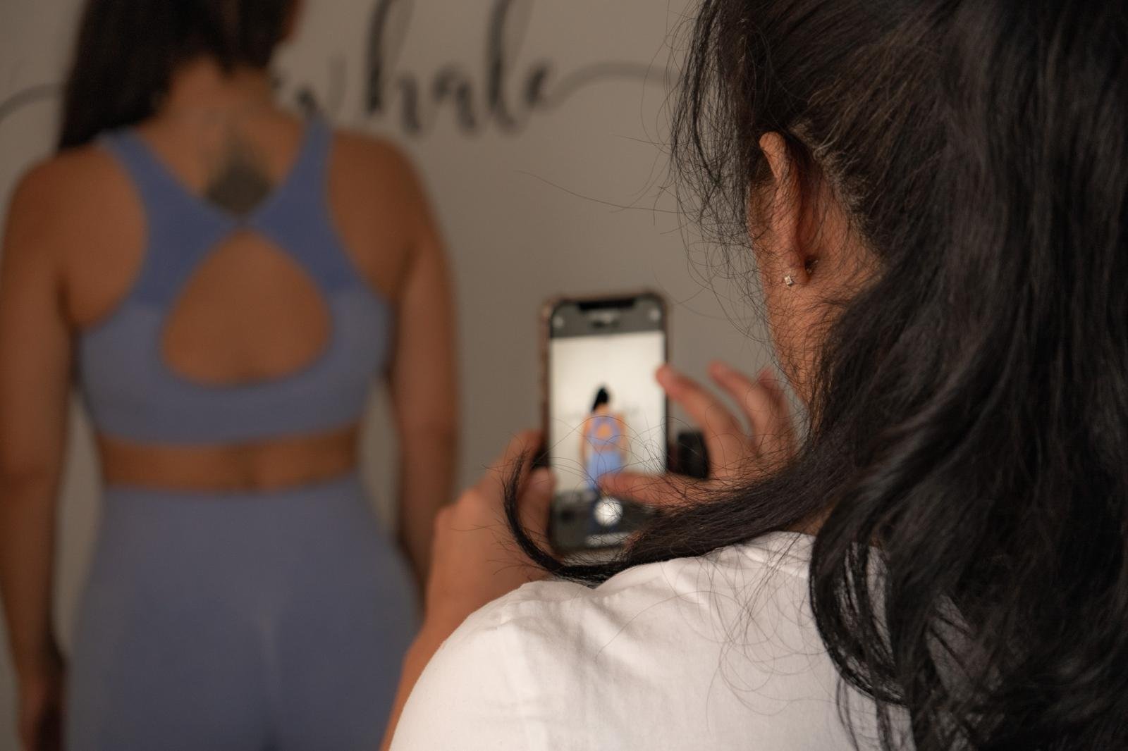 A woman taking a photo of another woman in front of a wall with writing on it, using her smartphone.