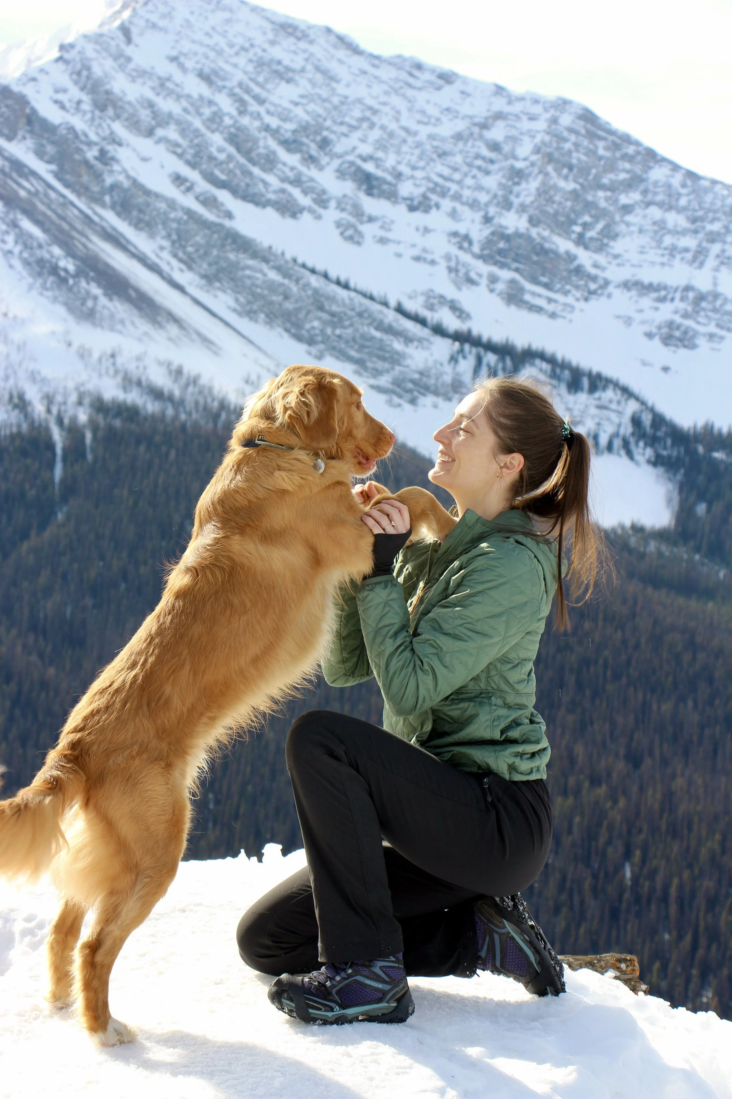 A woman therapist kneeling on snow in front of a mountain, smiling at a large duck tolling retriever dog that is standing on its hind legs, facing her, in a snowy mountain landscape.