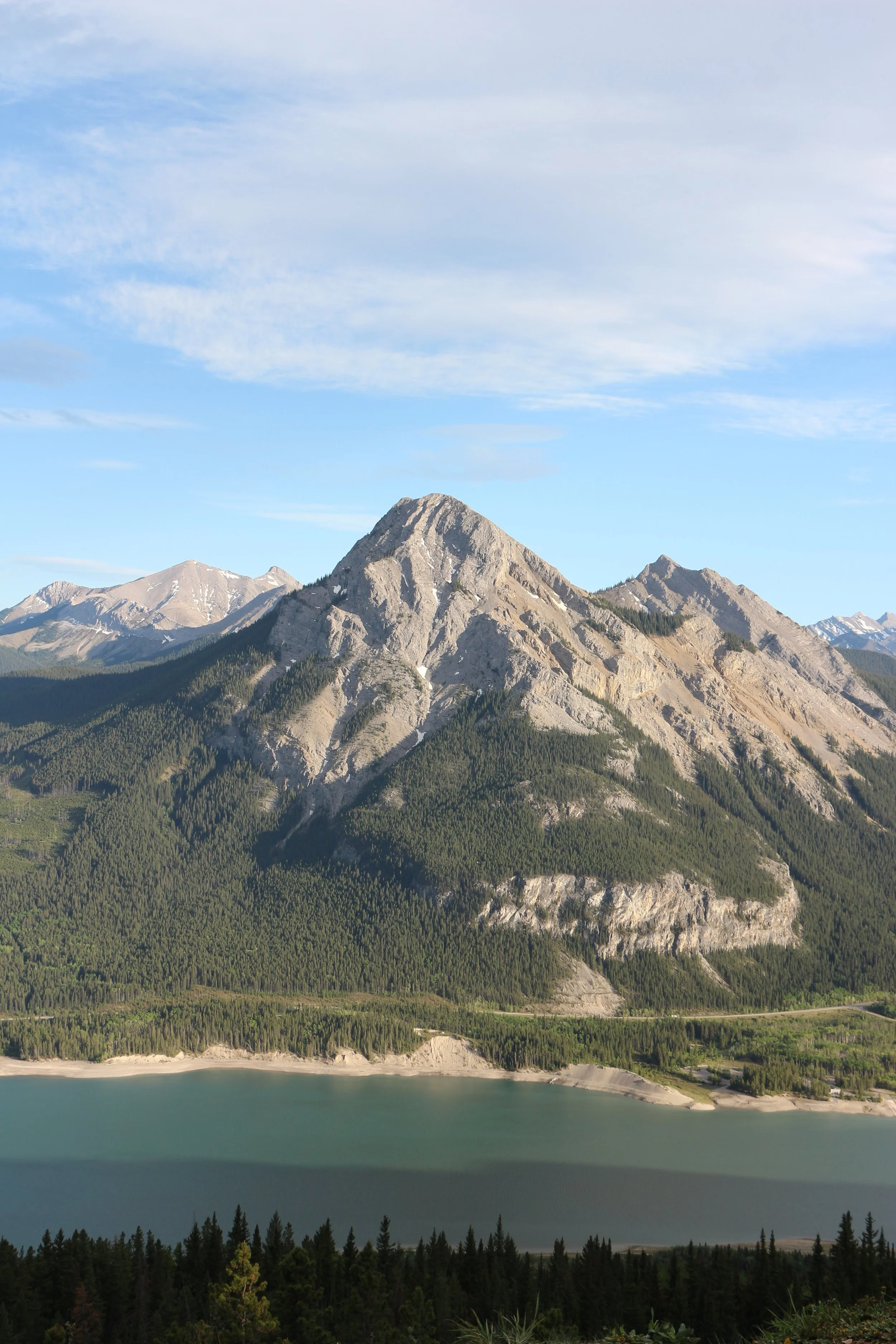 Photo of a large mountain with a forested base, a lake in the foreground, and a partly cloudy sky above.